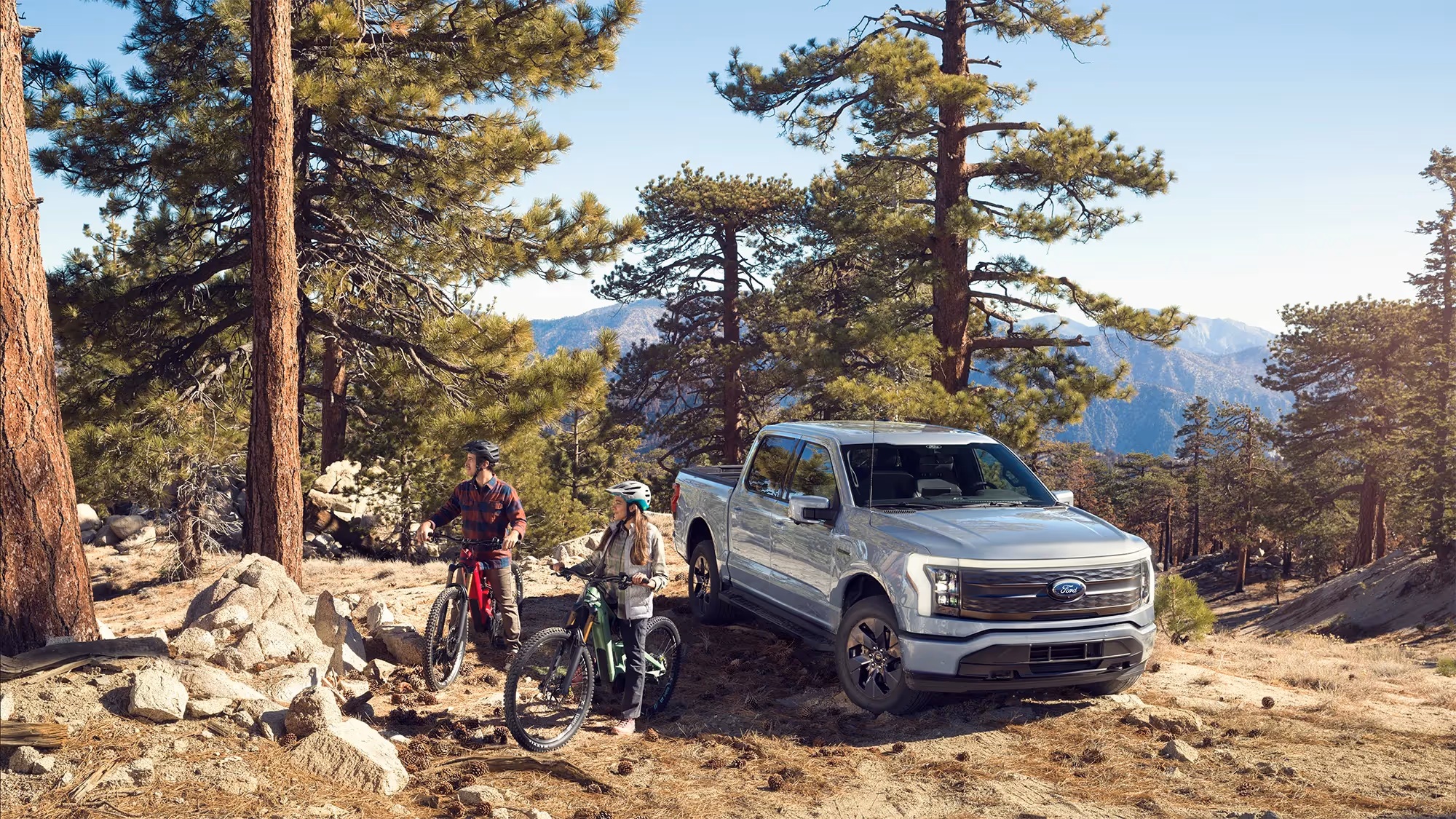 Two mountain bikers rest beside a silver Ford F-150 Lightning parked in a pine forest, sunlight streaming through the trees.