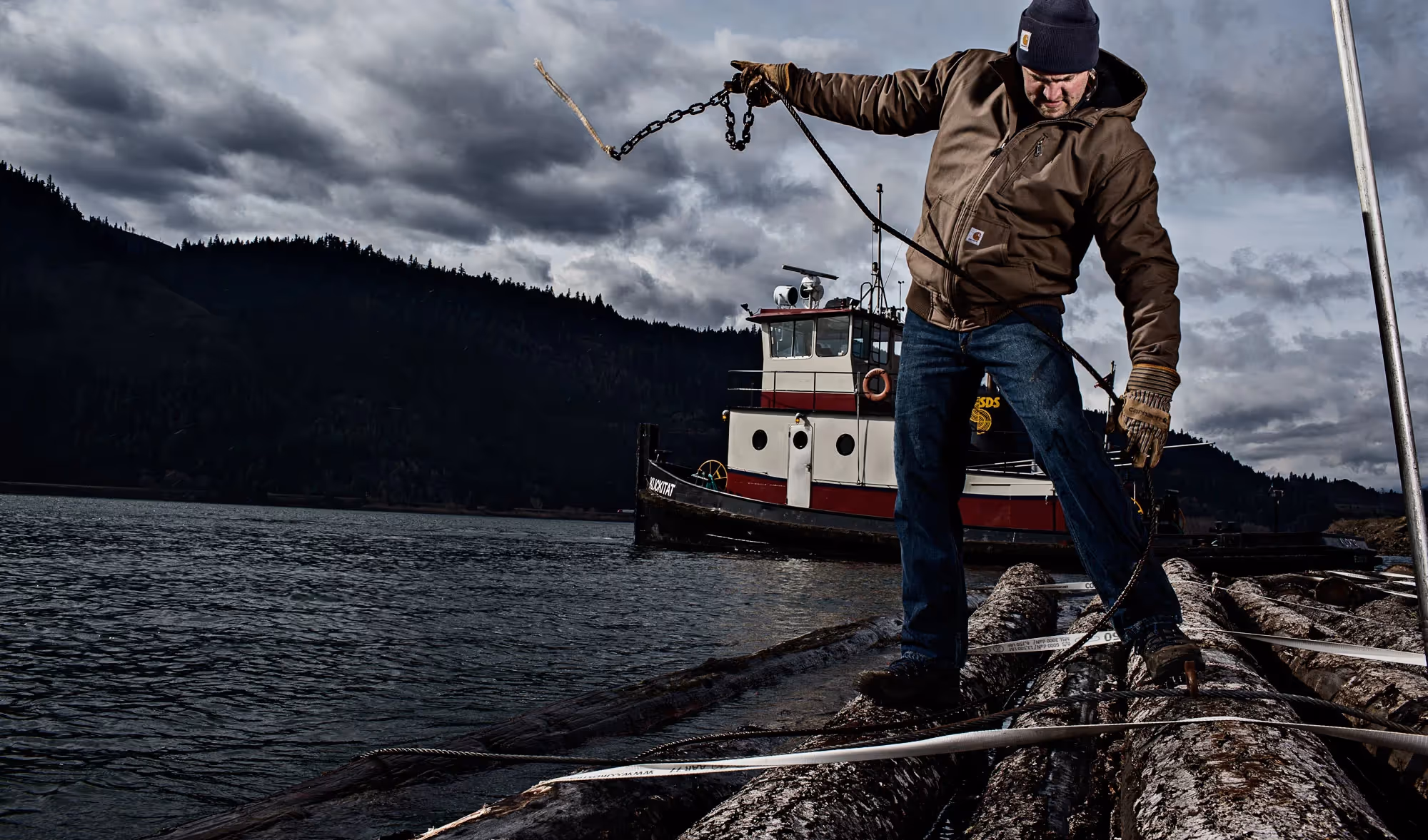 A man throws a chain to secure a log raft on a river, dark mountains and overcast skies behind him.