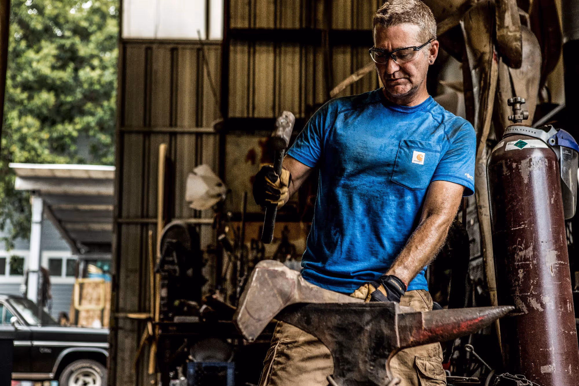 A blacksmith in a blue shirt works a piece of metal on an anvil inside a workshop, sparks flying as he strikes with a hammer.