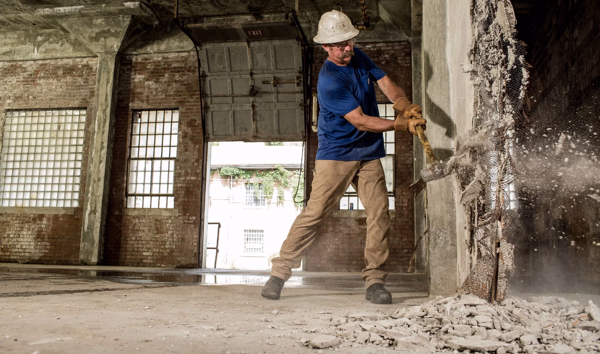 A demolition worker uses a sledgehammer to break through a crumbling wall inside an old industrial building.
