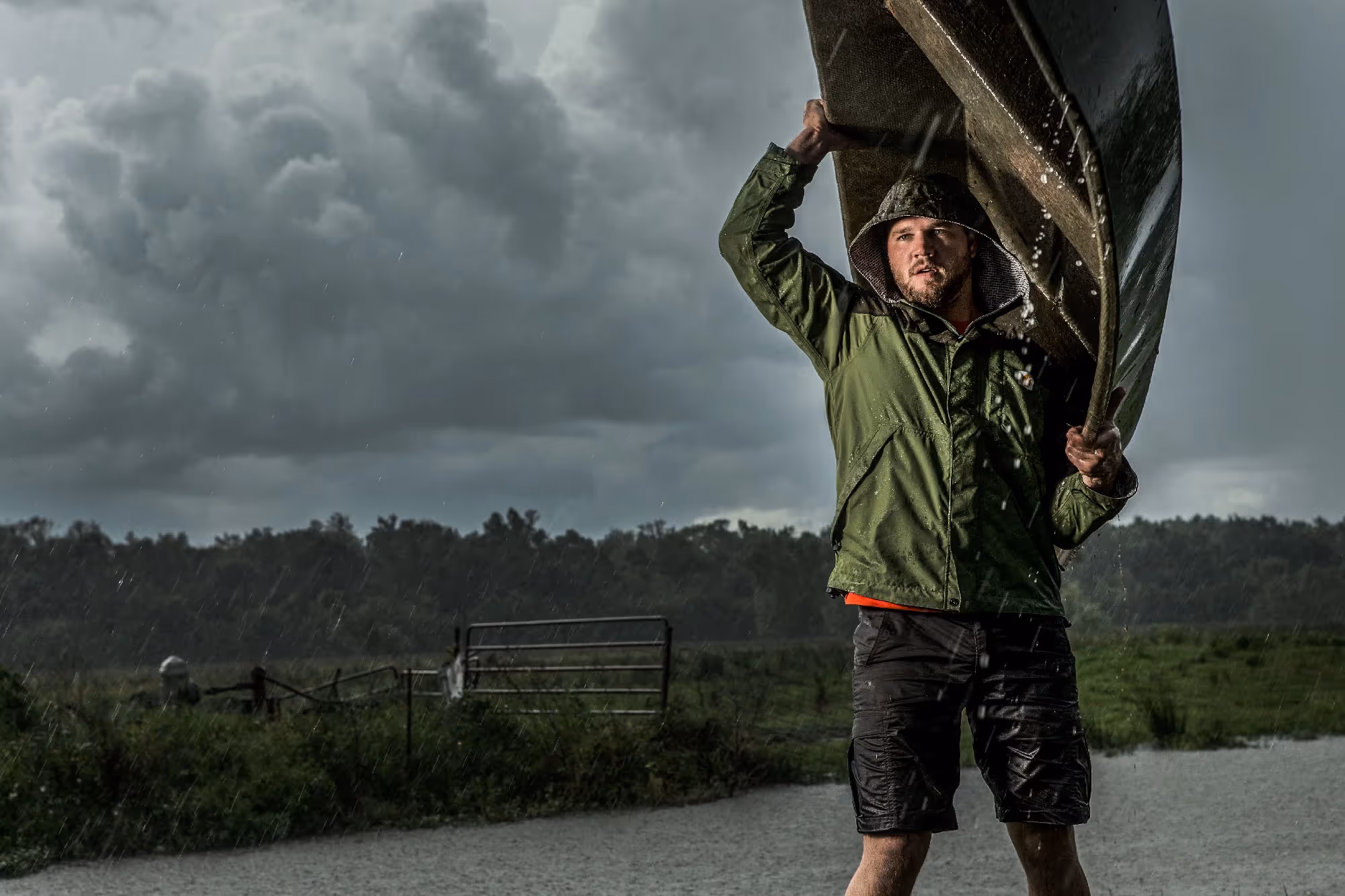 A man walks through heavy rain carrying a canoe on his shoulders, dark storm clouds rolling over a distant treeline.