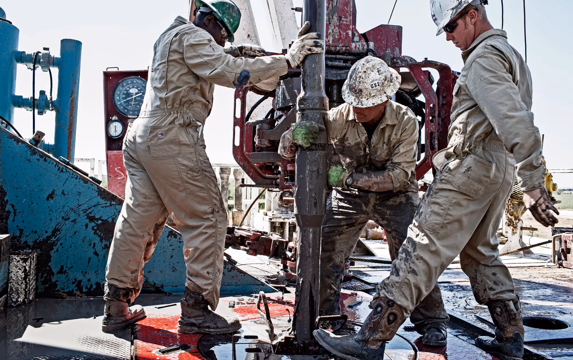 Three oil rig workers in tan coveralls operate heavy drilling machinery on an active job site.