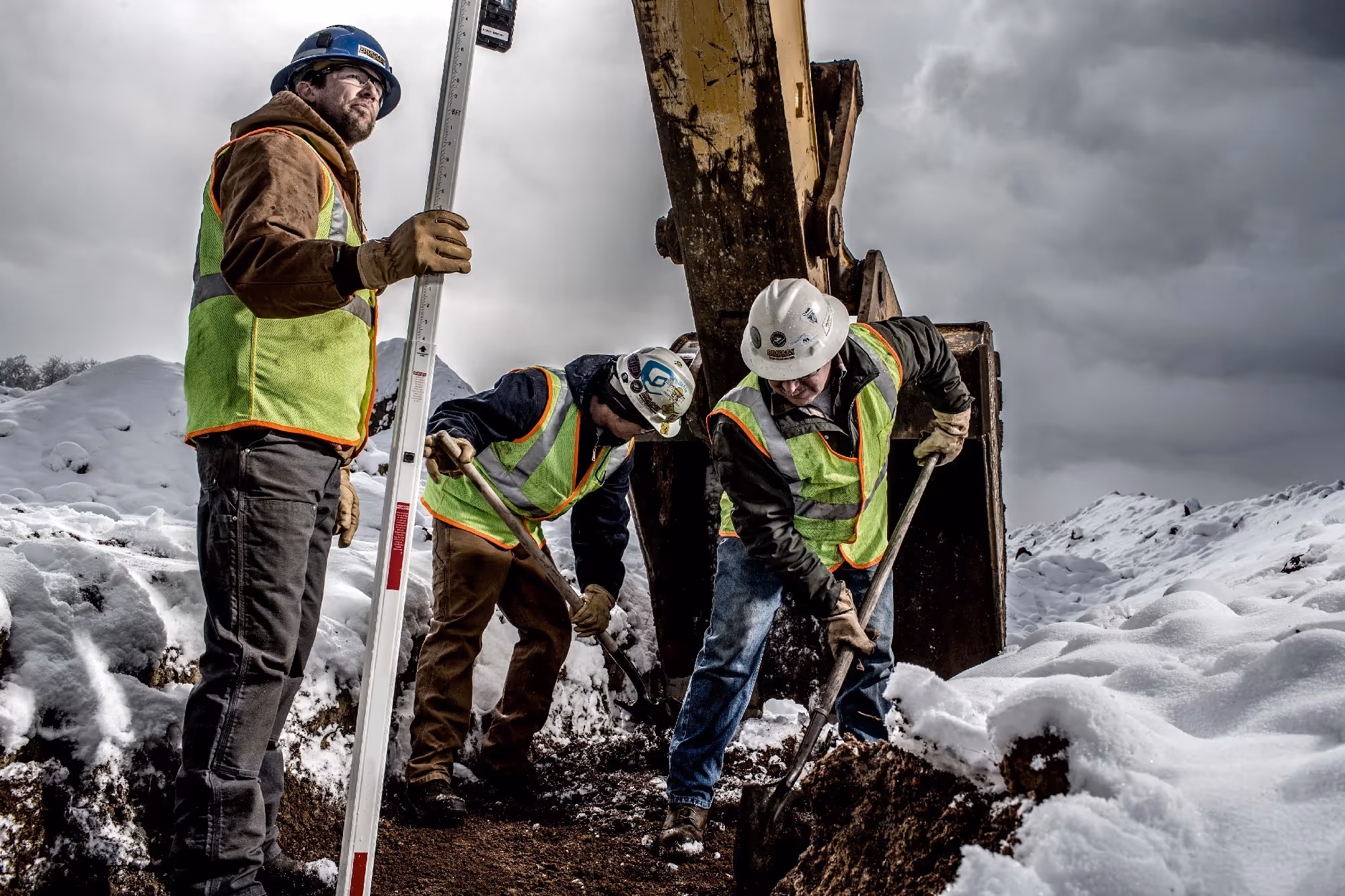 Construction workers in Carhartt gear measure ground levels near heavy equipment on a snowy mountain worksite.