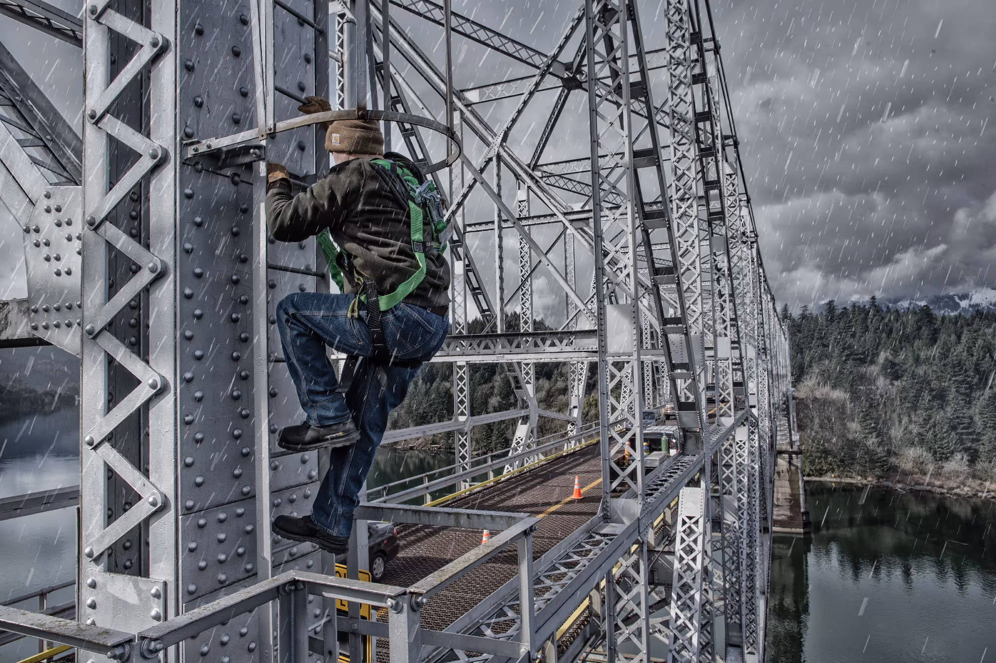 A construction worker climbs a steel bridge structure high above a river on a snowy day, safety harness strapped in place.