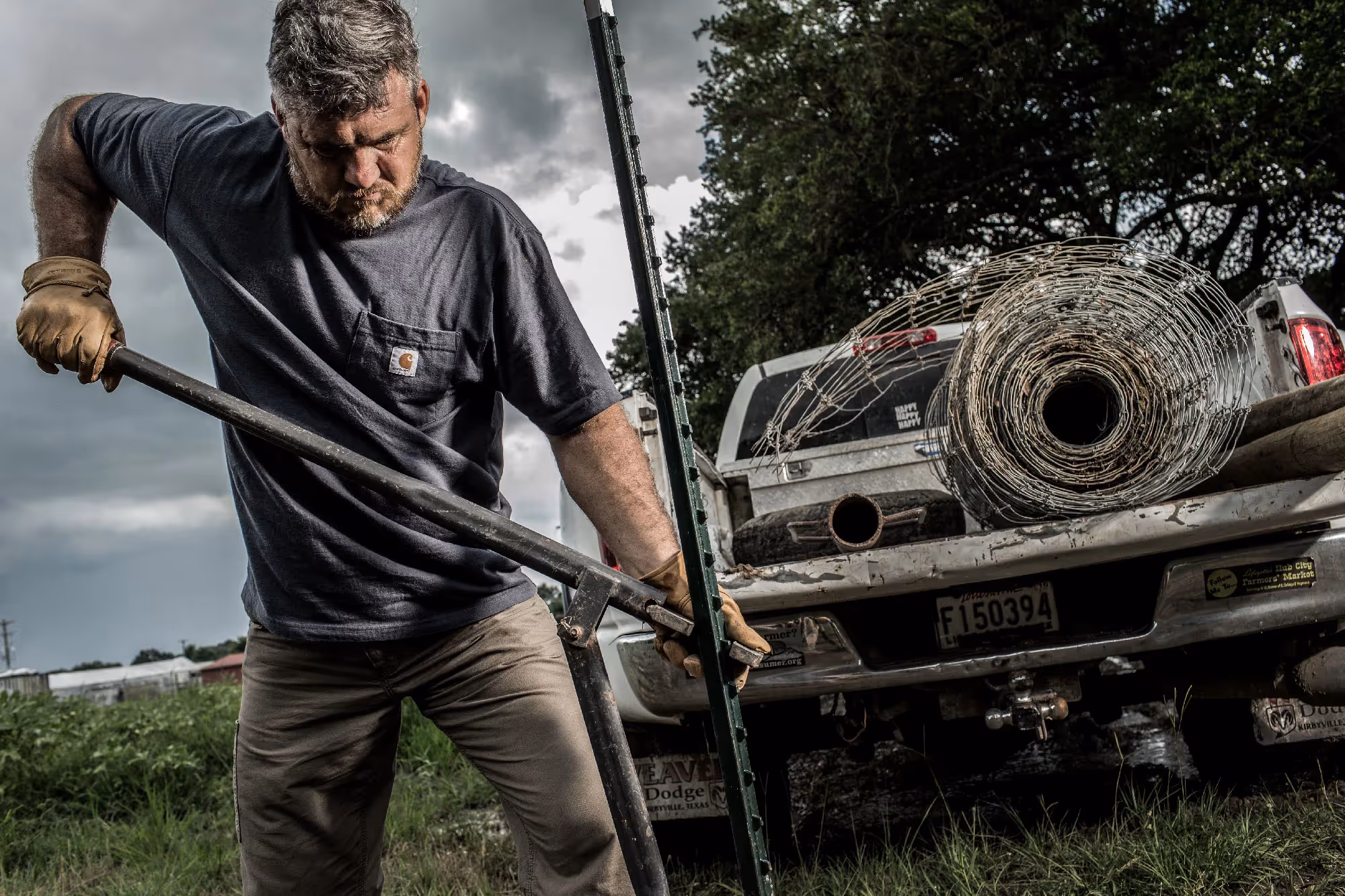 A man drives a steel fence post into the ground with a heavy mallet beside a pickup loaded with wire fencing.
