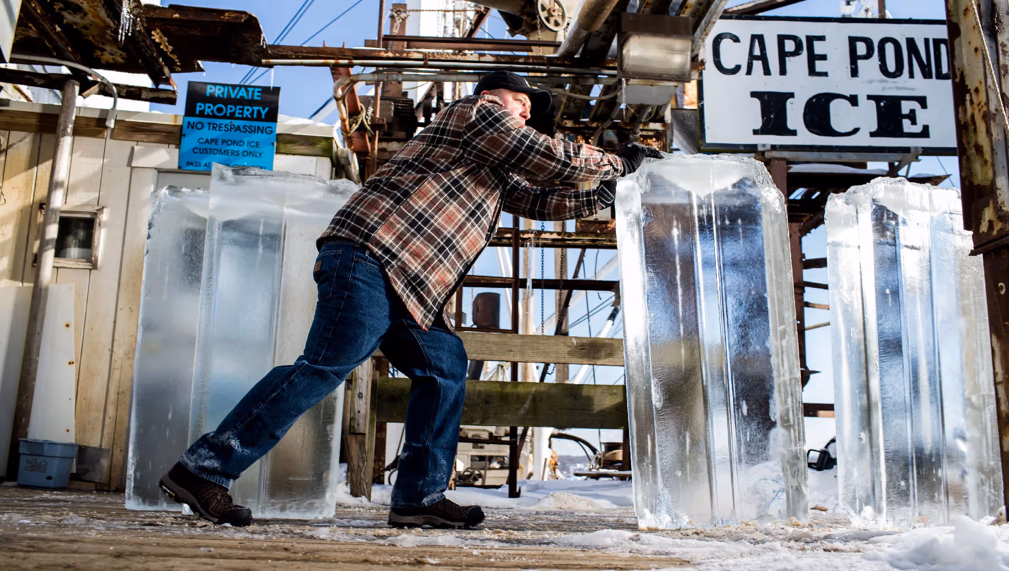 A man in a flannel shirt moves a massive ice block outside a facility marked “Cape Pond Ice,” sunlight reflecting off the frozen surface.