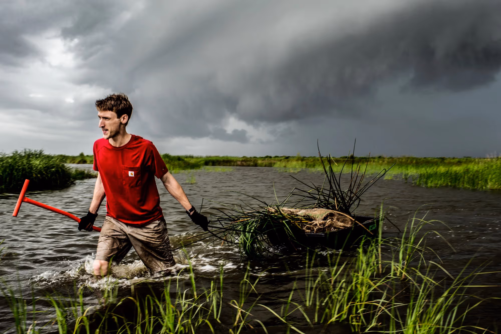 A man wades through waist-deep marsh water carrying a rake and net, storm clouds building in the distance.