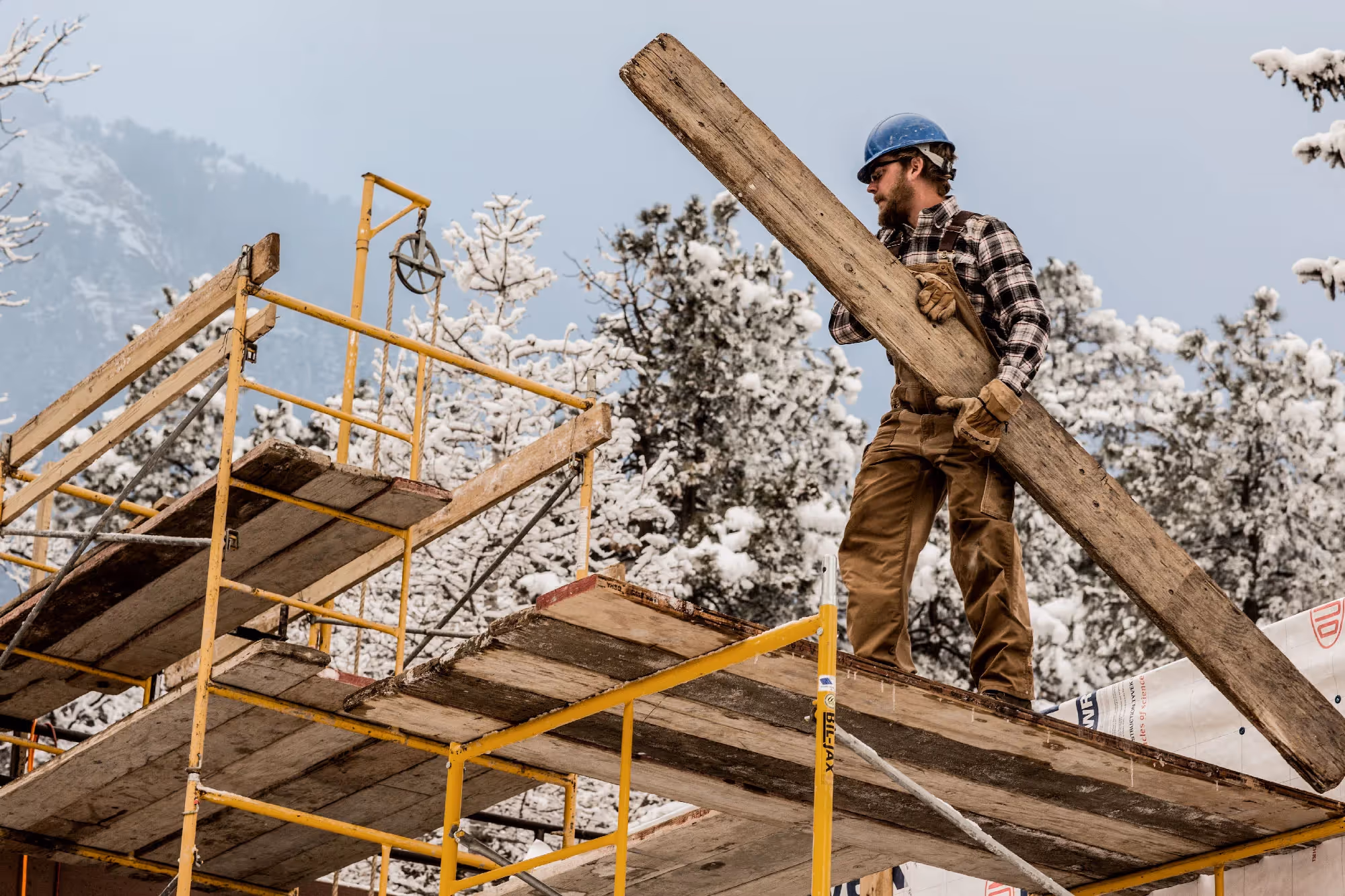 A construction worker in a hard hat carries a large wooden beam up scaffolding surrounded by snow-dusted pine trees.
