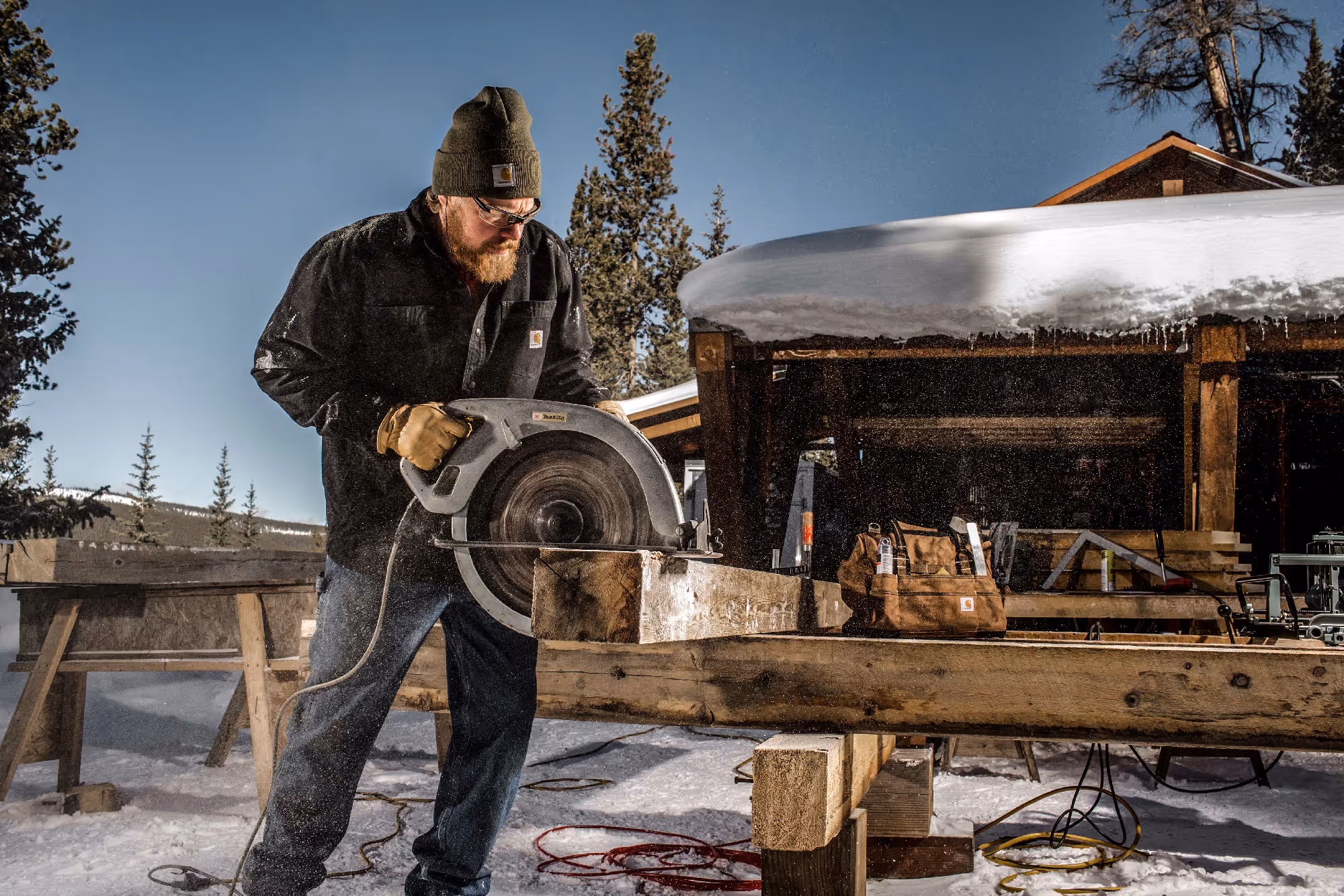 A carpenter uses a circular saw to cut lumber outside a cabin under a clear winter sky, snow covering the ground around him.