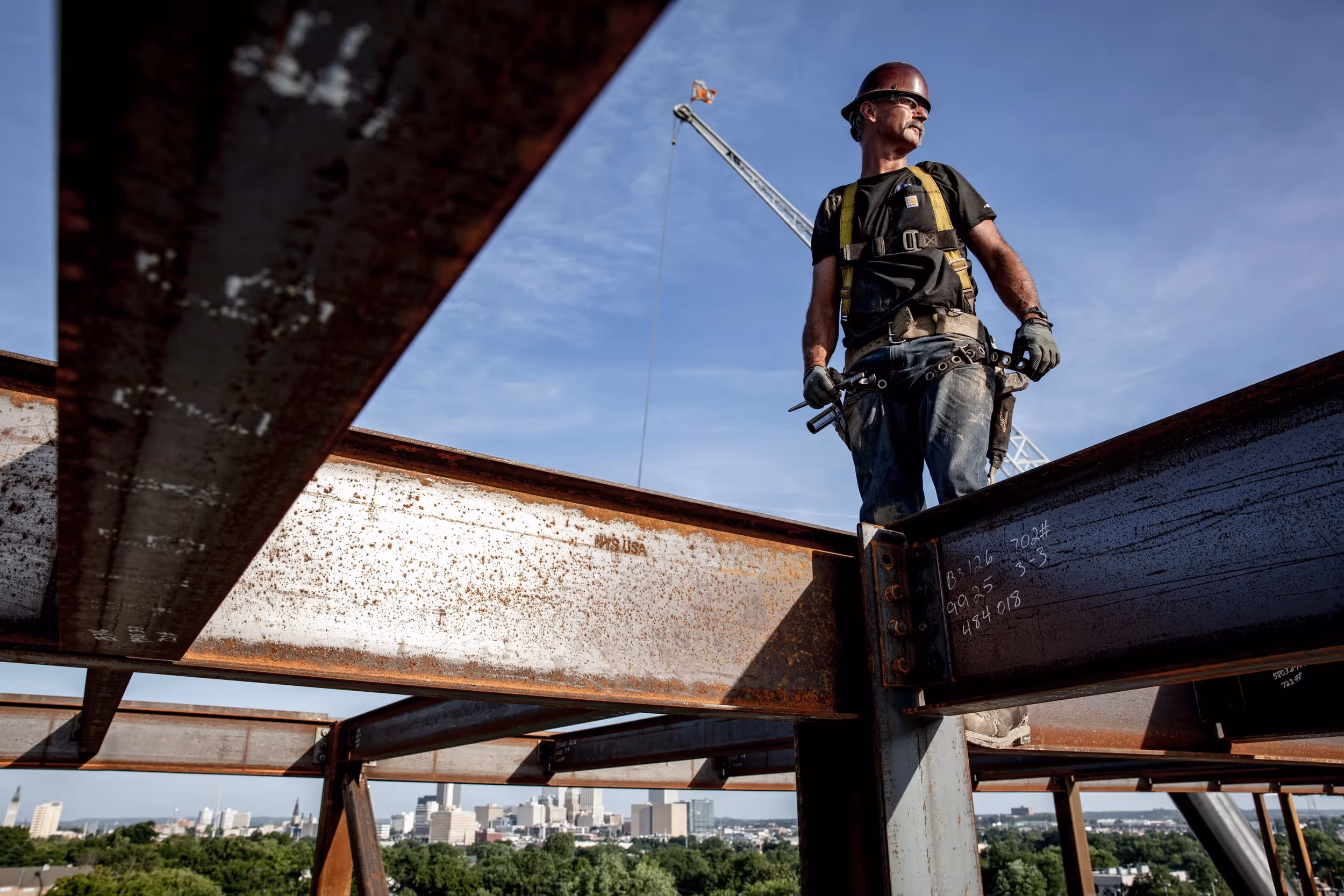 An ironworker in a safety harness walks across a steel beam high above the city skyline, sunlight hitting the metal structure.