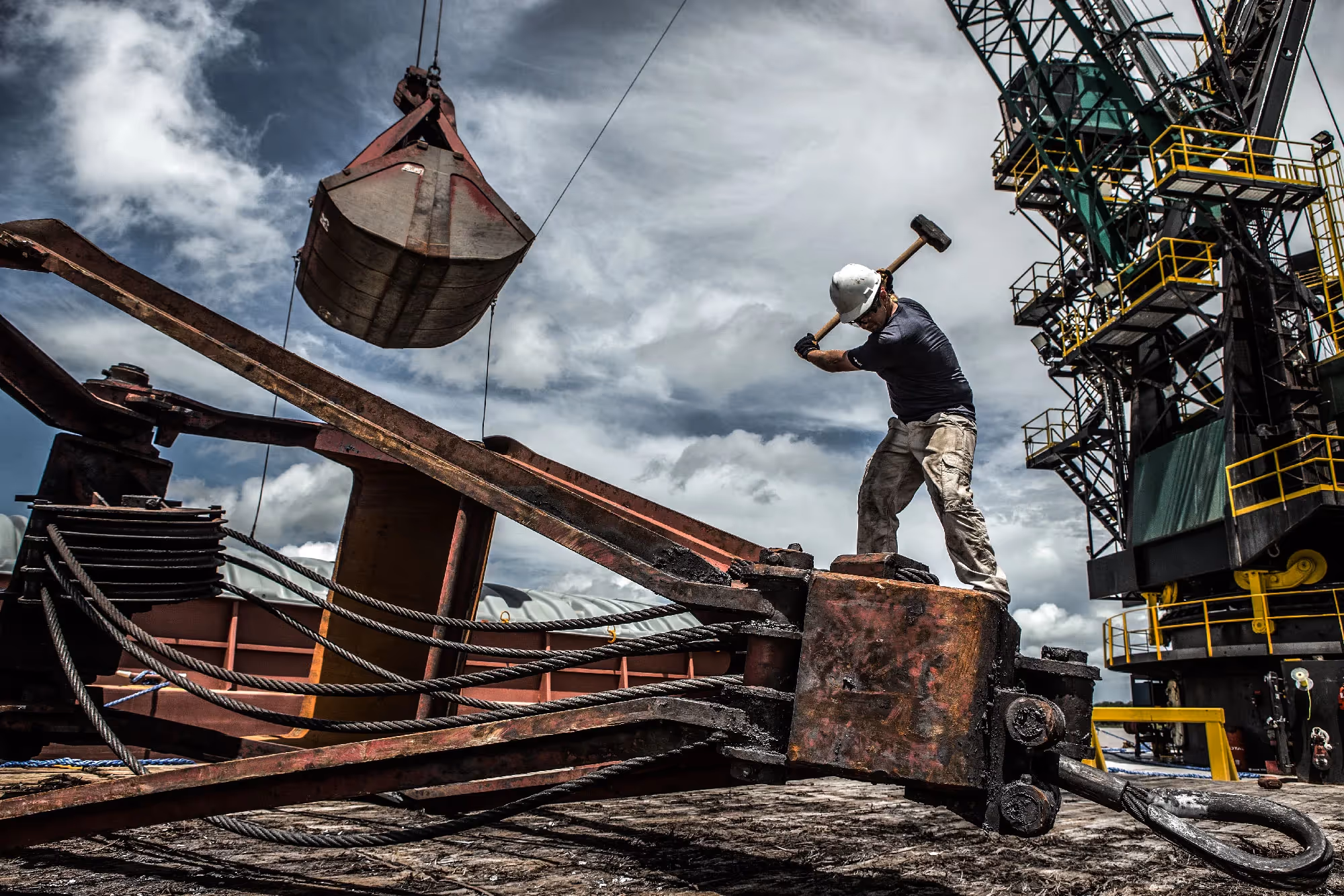 Two workers guide a suspended load from a crane on a ship deck, one man using a sledgehammer to adjust the rigging.