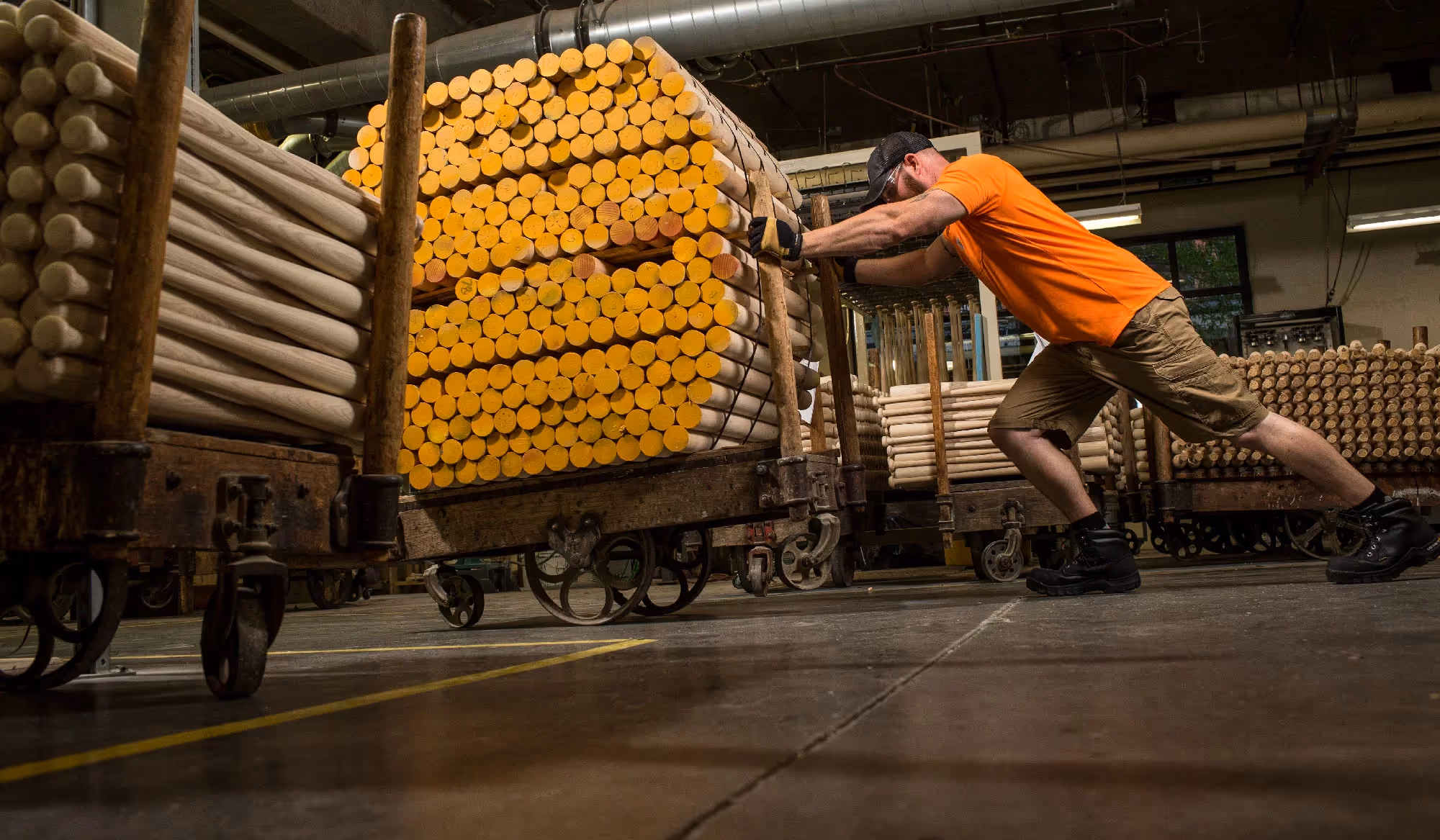 A worker in an orange shirt pushes a large cart loaded with stacked industrial materials through a factory floor.