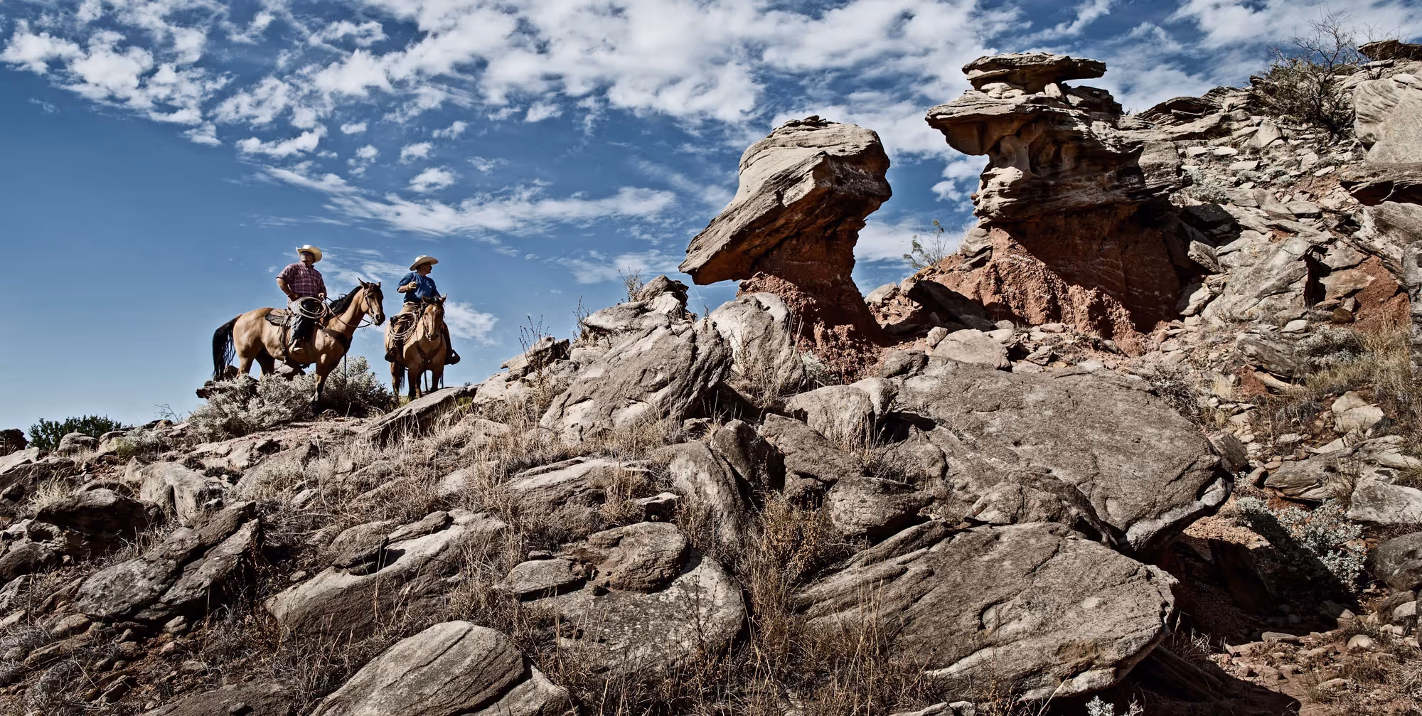 Two riders on horseback navigate rocky terrain under a wide blue sky, desert cliffs and formations in the background.