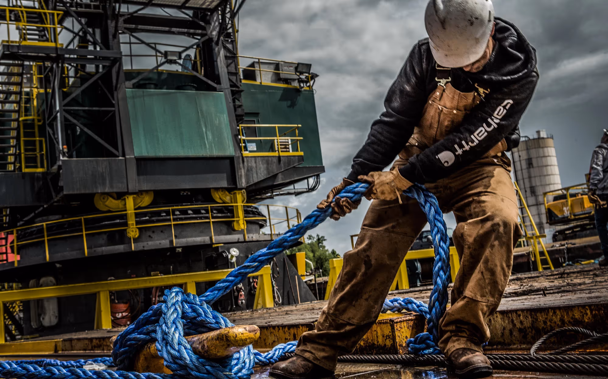 A worker in a hard hat and gloves hauls a heavy blue rope across a dock at an industrial site, machinery and cranes behind him.