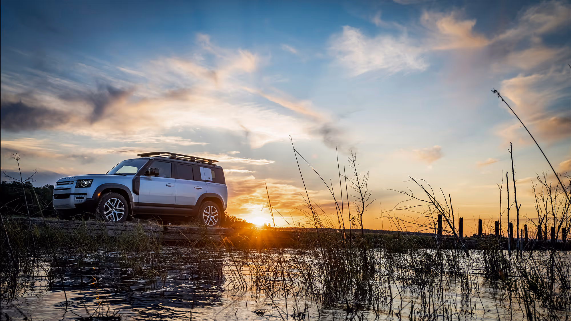 The Defender parked near tall reeds by the water’s edge at sunset, sky painted with orange and blue tones.