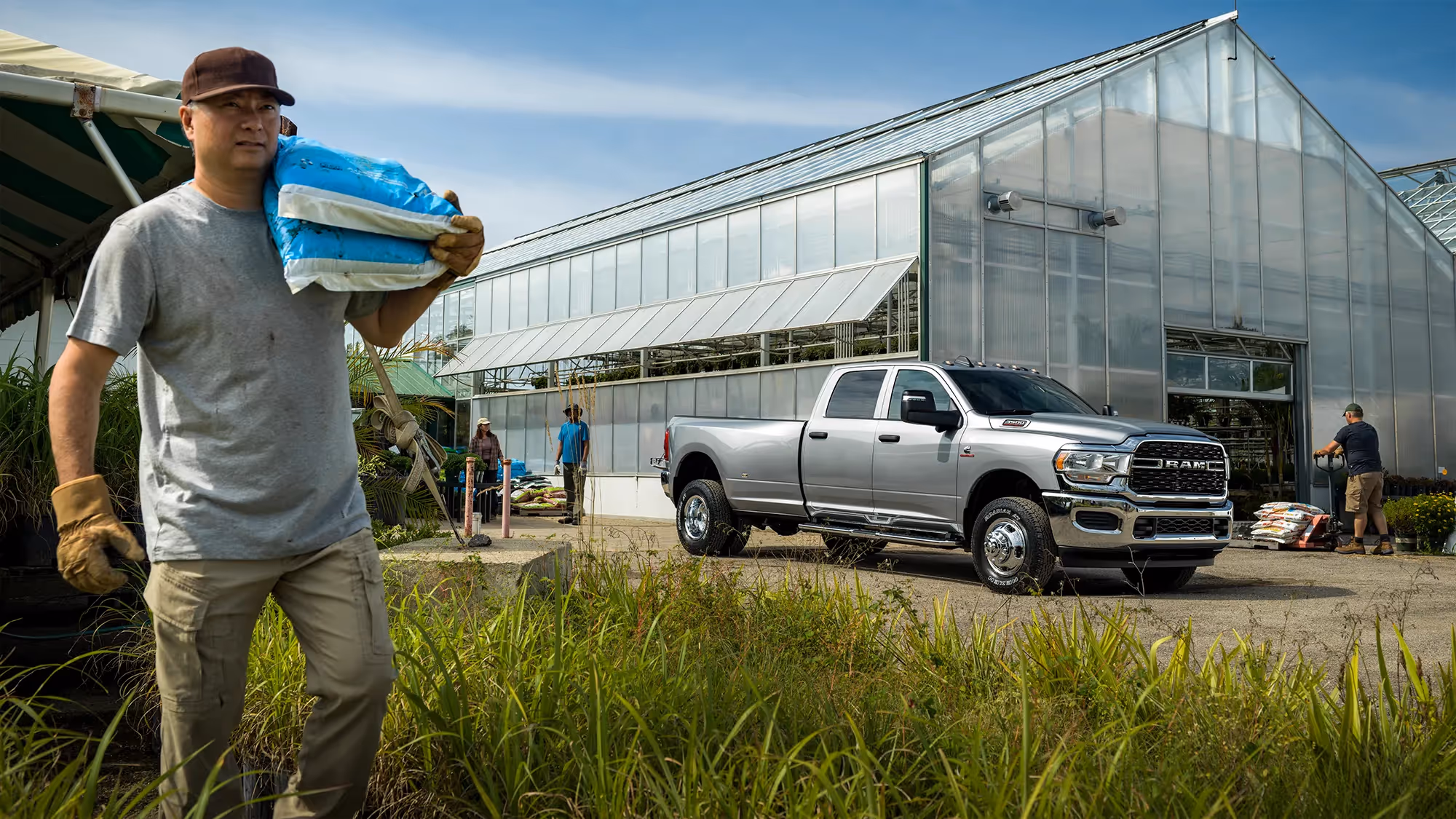 A man carries stacked trays of plants across a field toward a Ram HD truck parked near the greenhouse.