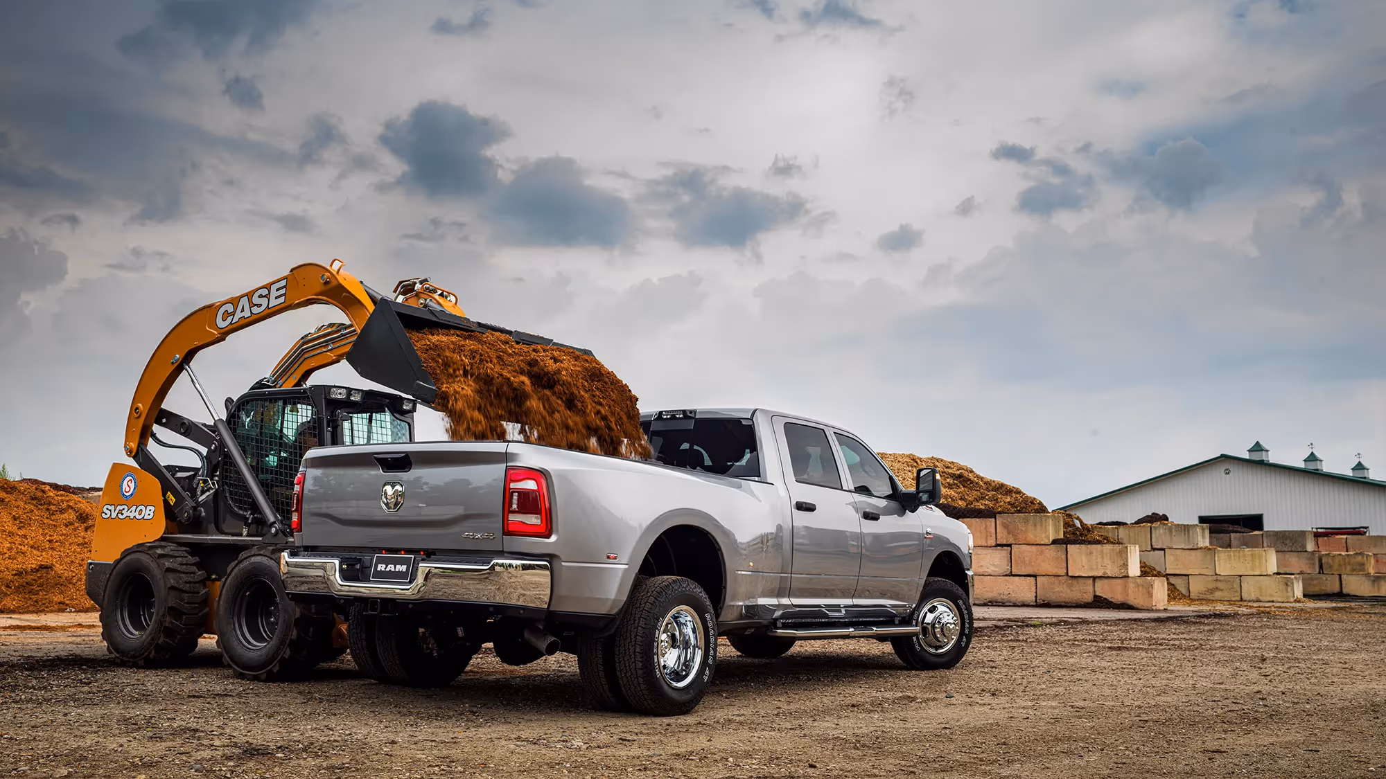 A silver Ram HD pickup loaded with soil parked beside an excavator at a job site.
