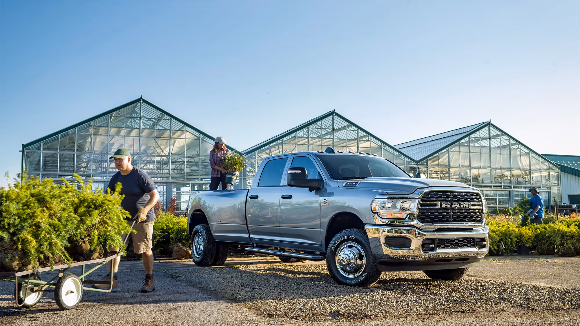 Workers load plants onto a trailer beside a Ram HD parked near glass greenhouses.