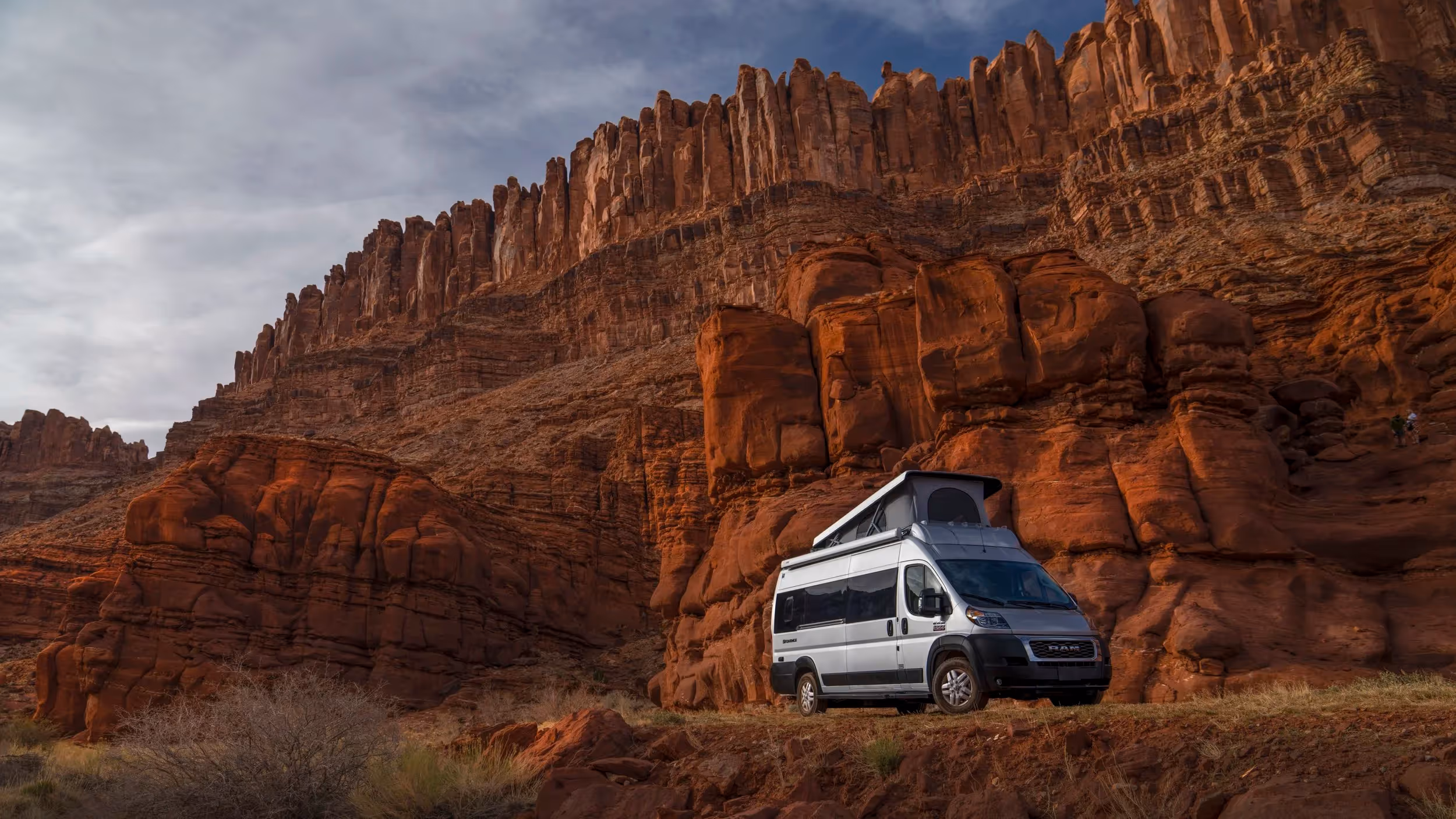 A Winnebago Revel framed against red desert cliffs under a bright blue sky.