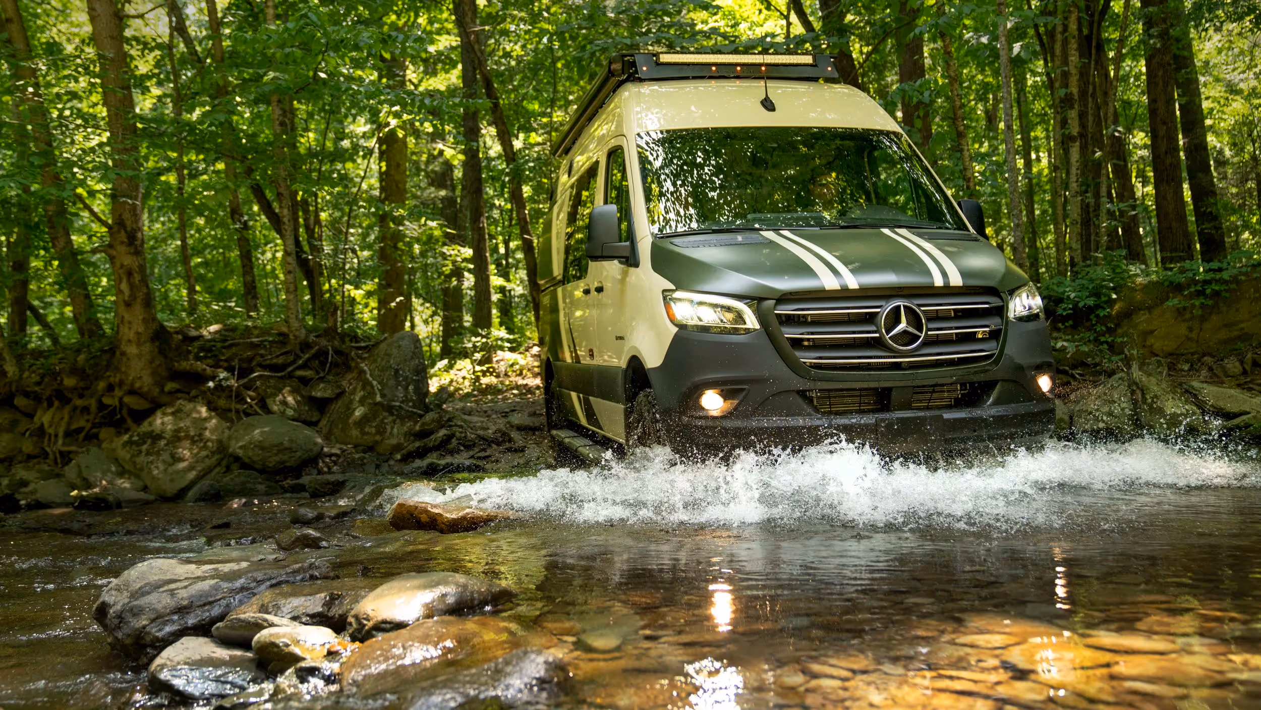 A white Winnebago Revel drives through a shallow rocky stream under a canopy of trees.