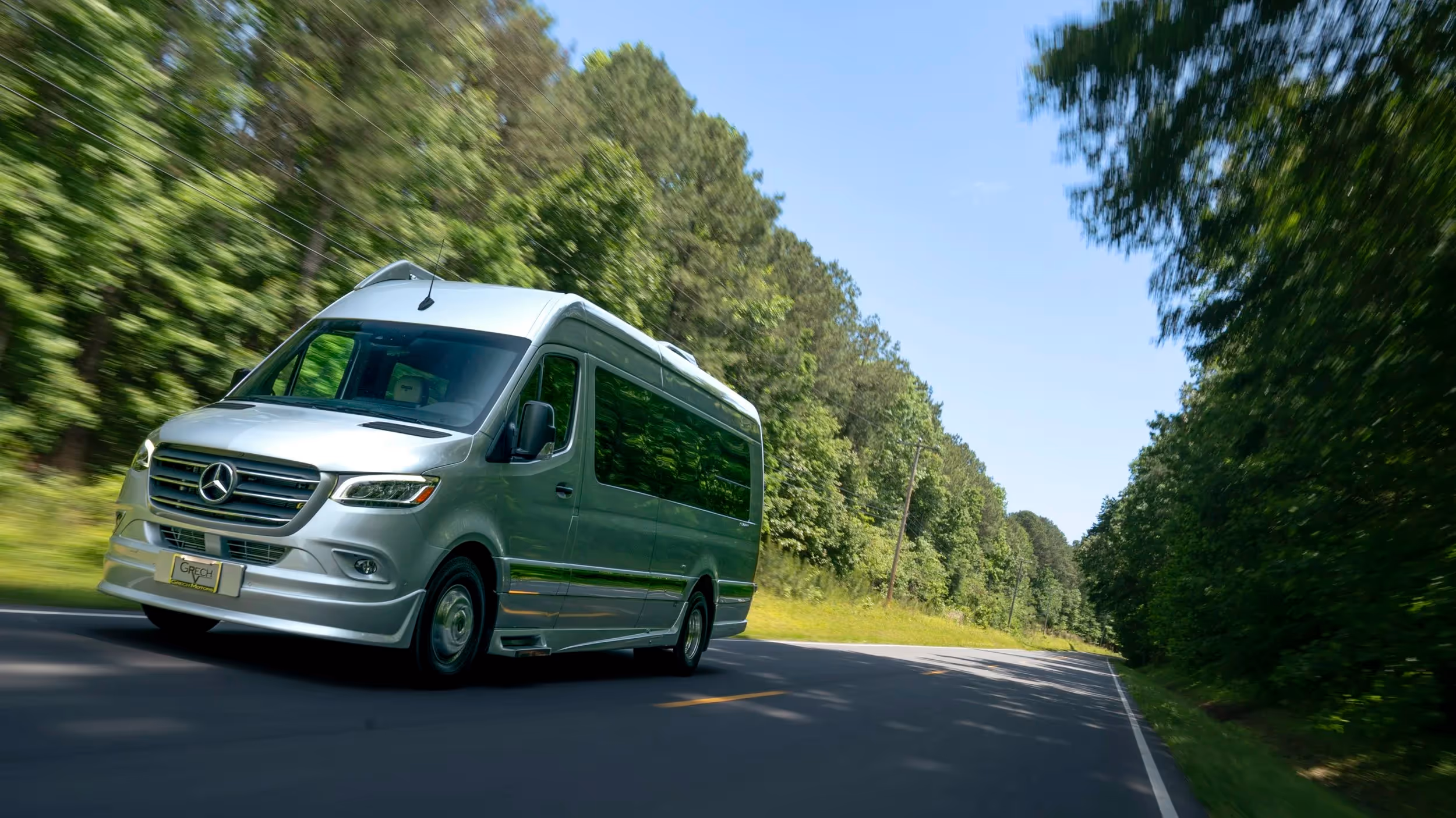 A silver Winnebago Revel drives down a forest highway, sunlight glinting off the body.