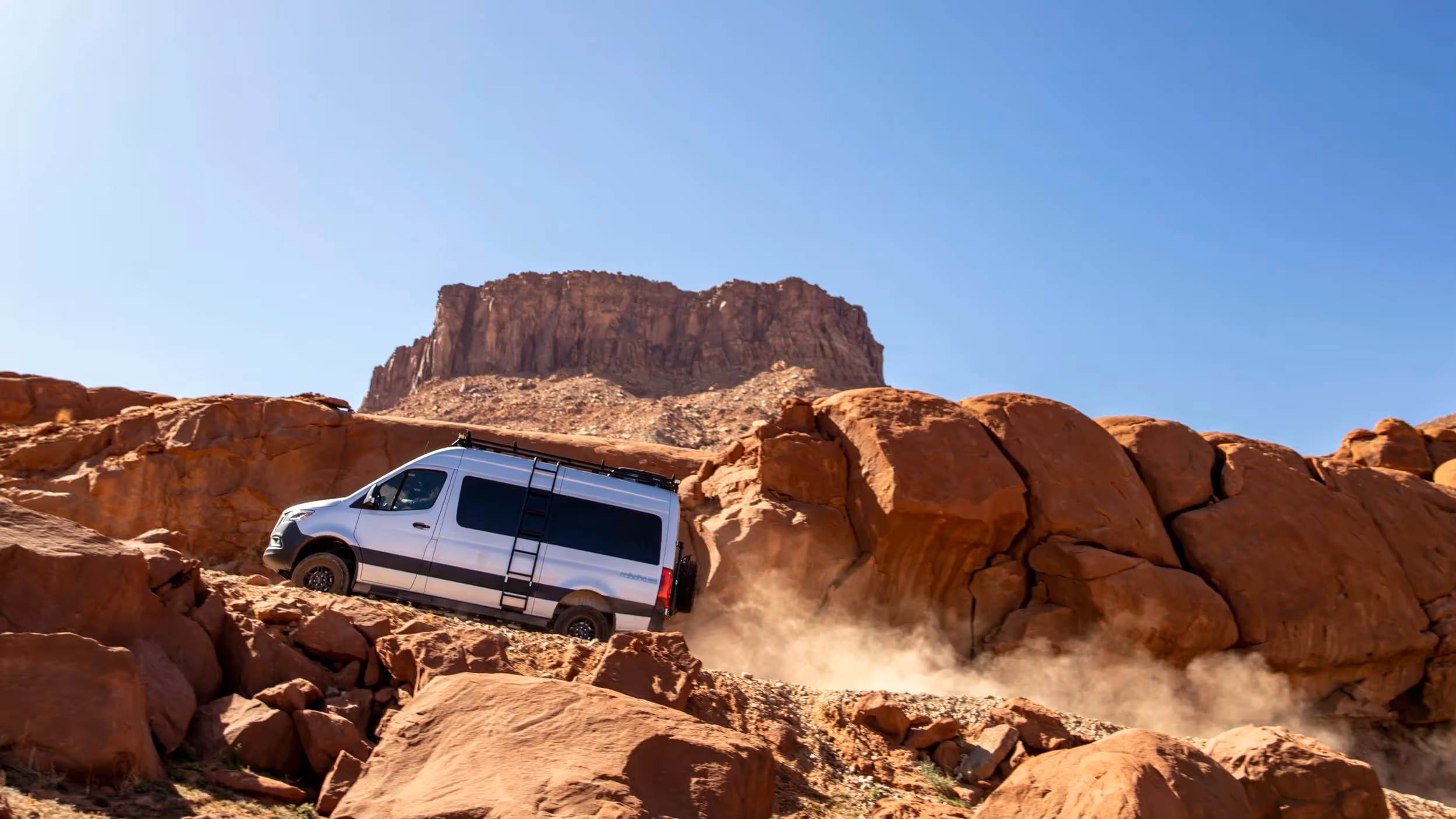 A Mercedes-Benz Sprinter 4x4 adventure van climbs through red desert rock terrain, kicking up dust beneath a bright blue sky.A Mercedes-Benz Sprinter 4x4 van powers uphill through rugged desert rocks and sand, sunlight cutting through the dust trail behind