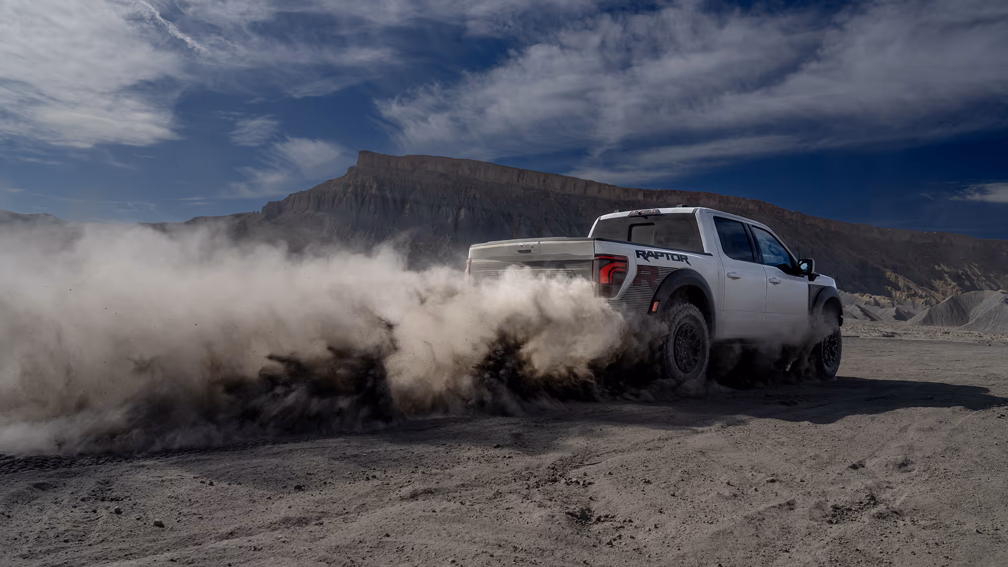 Rear view of Ford Raptor R sliding through dirt with heavy dust cloud in rocky landscape