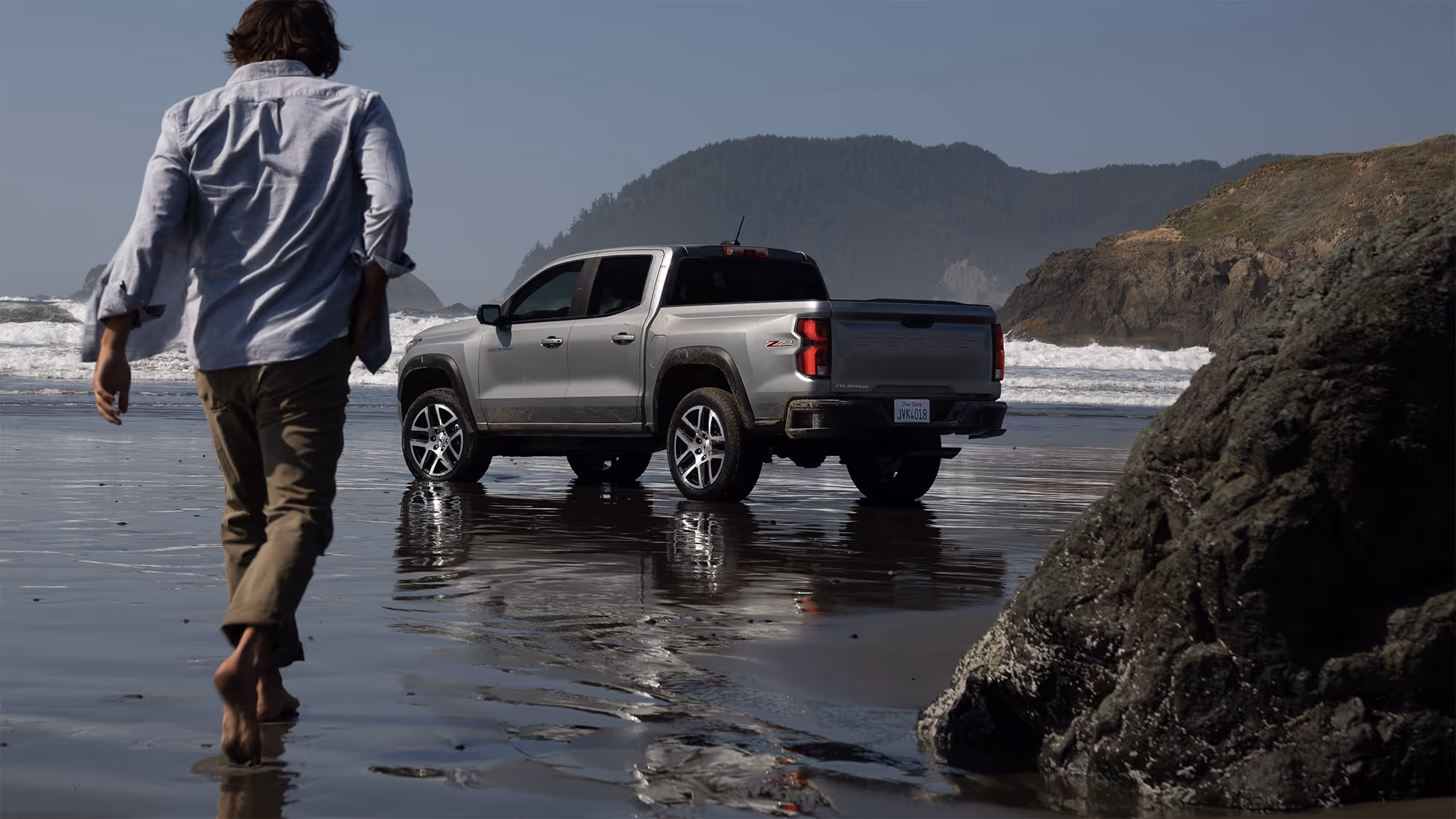 Man walking barefoot toward a Chevrolet Colorado ZR2 parked on wet sand with waves and coastal cliffs