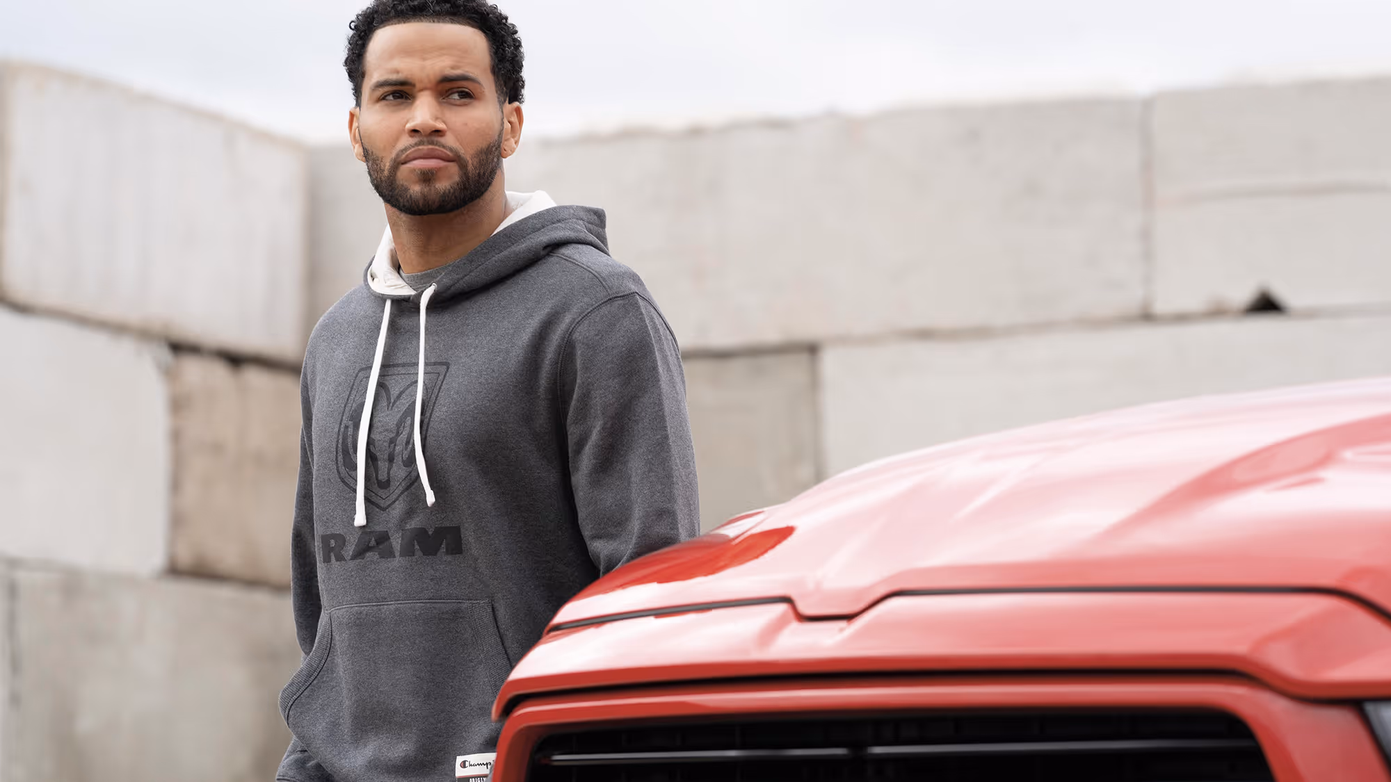 Man in RAM hoodie standing beside a red truck in an industrial setting, conveying strength and everyday utility.
