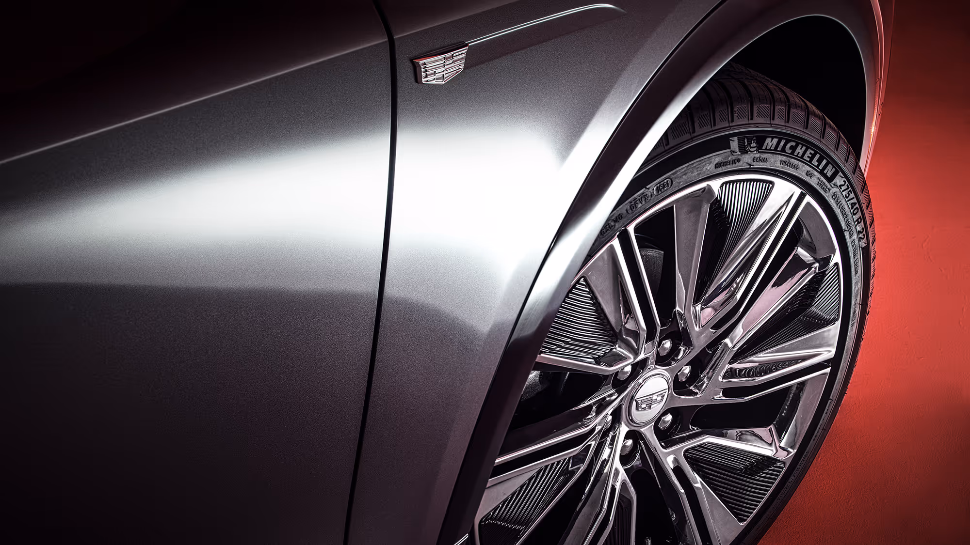 Close-up of Cadillac LYRIQ front wheel and fender, showcasing metallic gray paint, chrome rim detail, and Michelin tire against a red gradient background.