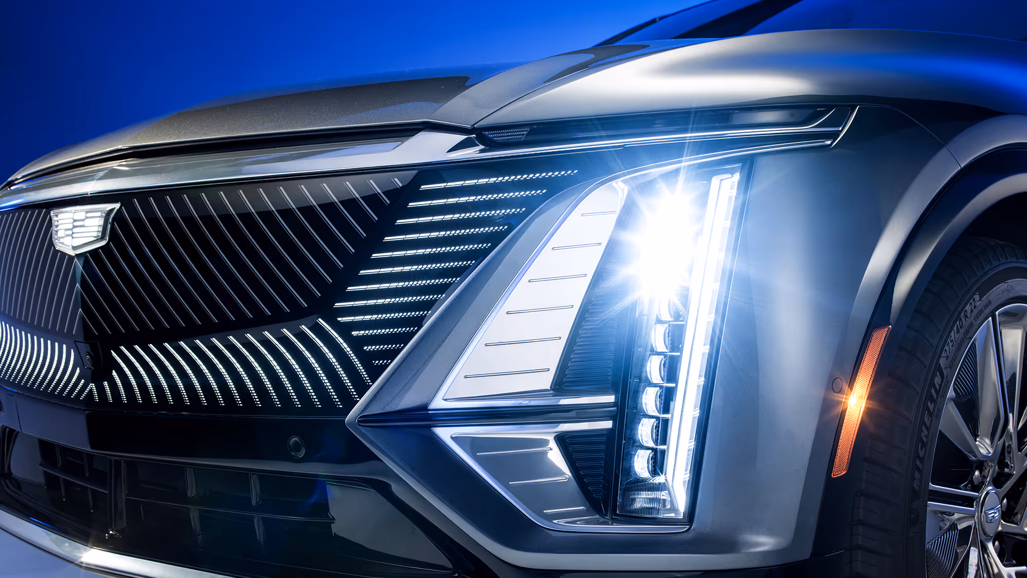 Close-up of Cadillac LYRIQ front grille and headlight with illuminated LED elements against a blue background.