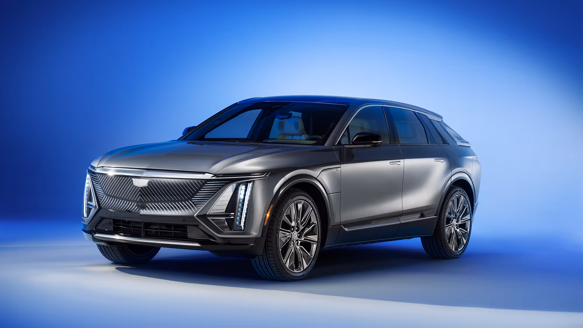 Front three-quarter view of Cadillac LYRIQ in metallic gray with illuminated grille and headlights, set against a blue gradient background.