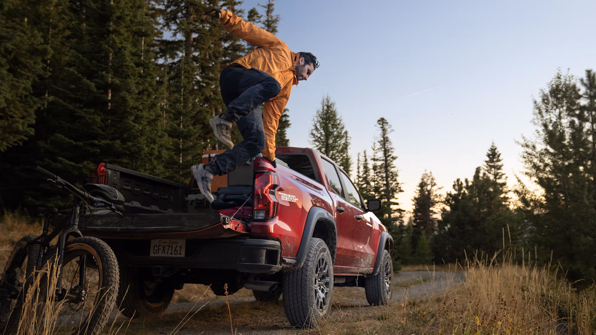 Man jumps down from the bed of a red Chevrolet Colorado Trail Boss parked off-road, with a mountain bike and gear visible in the truck bed.