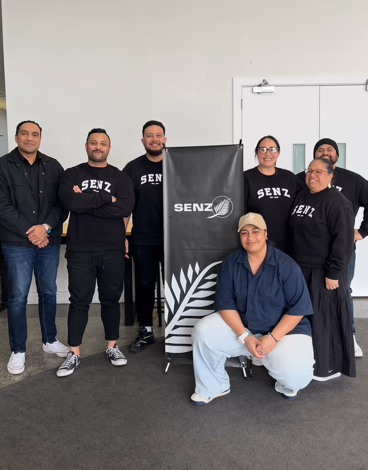 Group of seven people posing indoors around a black banner with the word SENZ and a fern leaf logo.