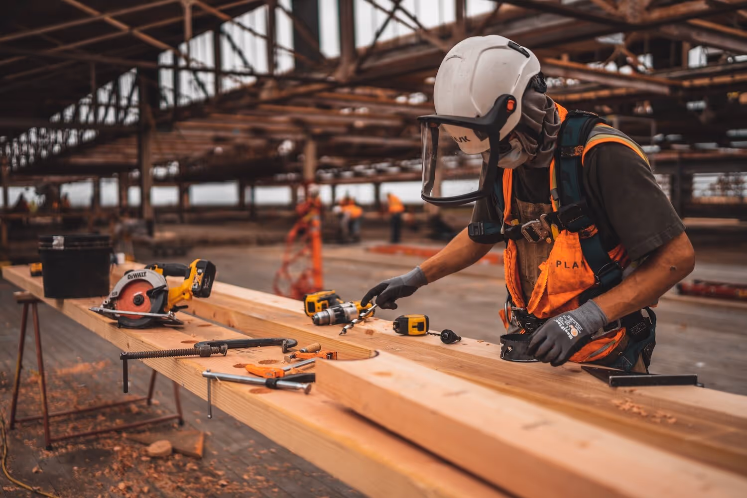 Construction worker wearing helmet and safety gear using a drill on wooden planks in an industrial setting.