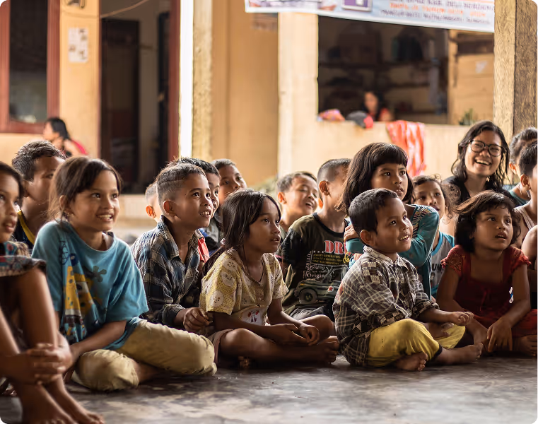 Group of attentive children sitting cross-legged on the floor indoors, watching something with interest and smiling.