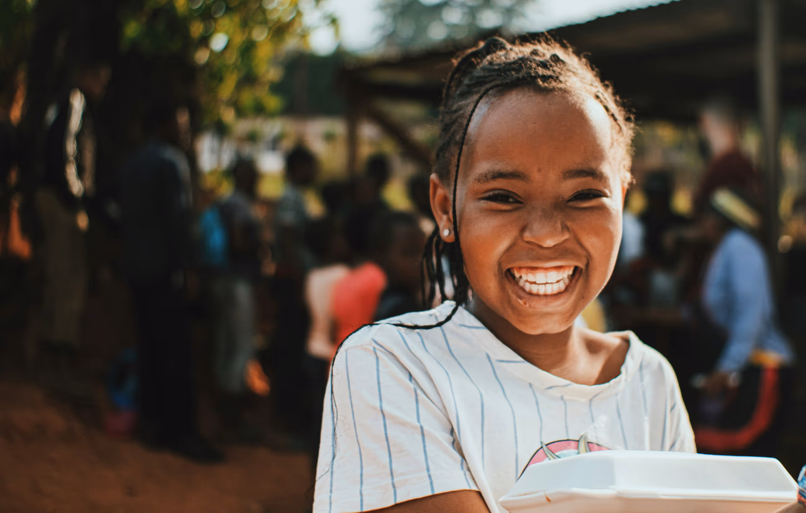 Smiling girl with braided hair holding a white food container outdoors with blurred people in the background.