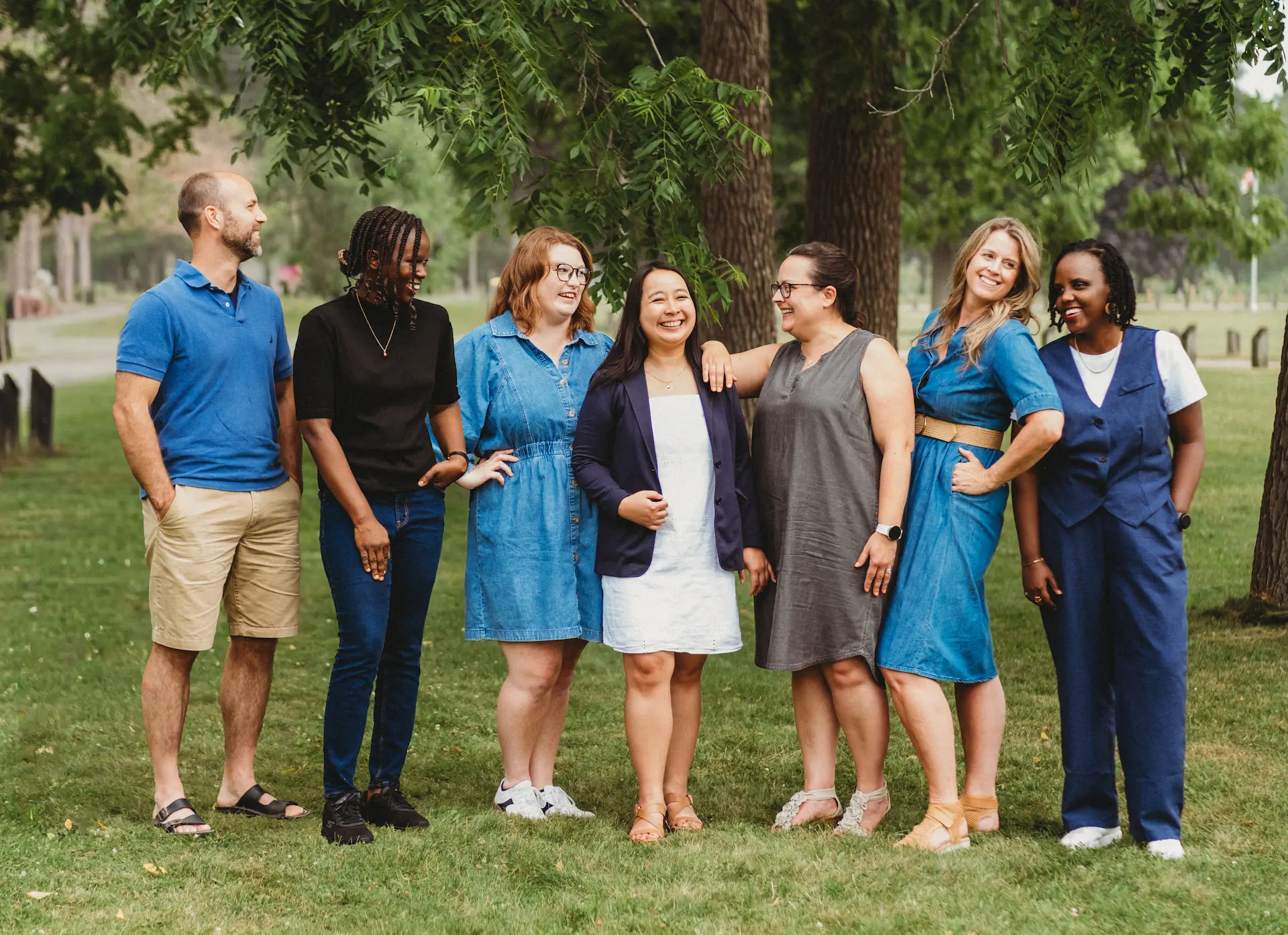Group of six women and one man standing on grass in a park, smiling and interacting under green trees.