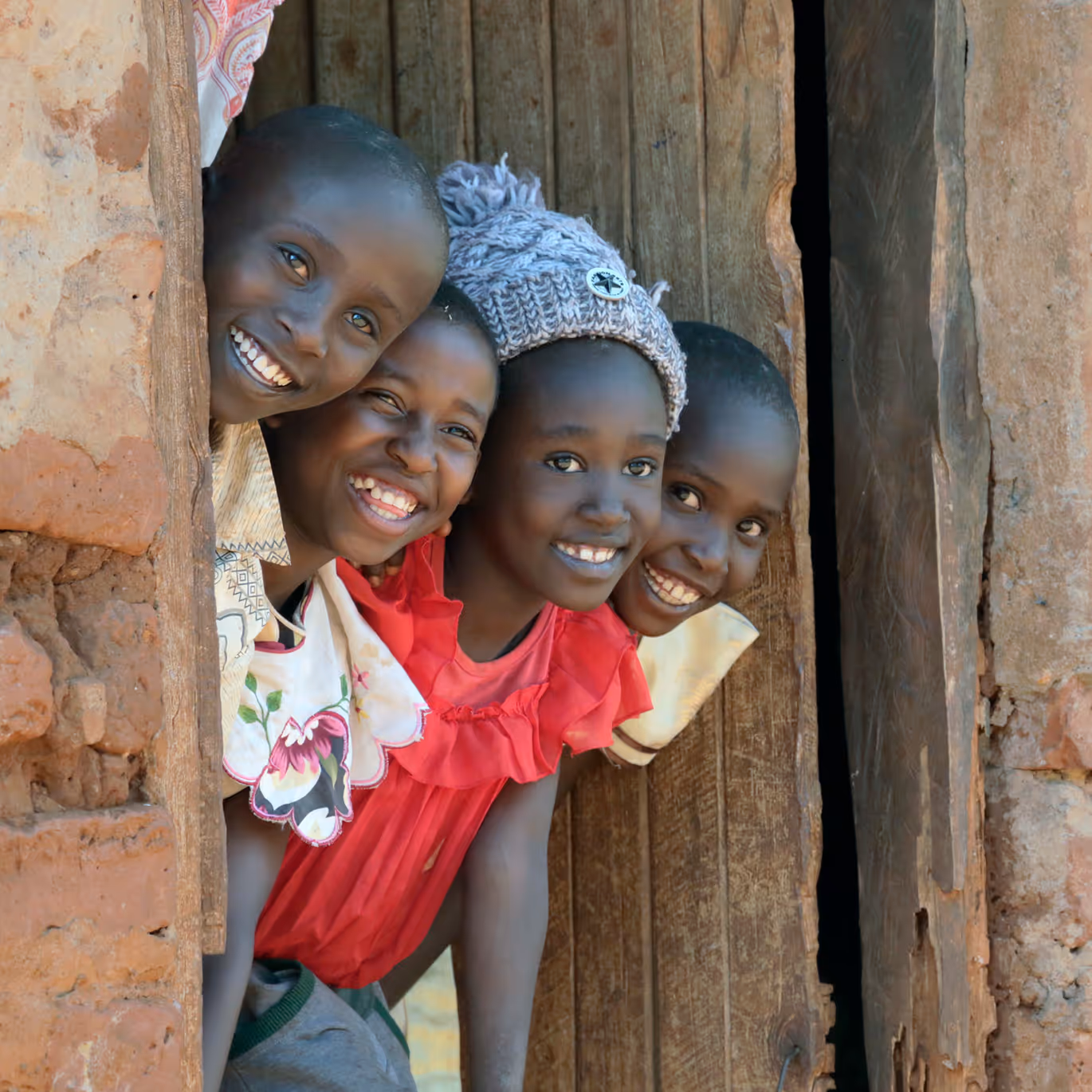Four smiling children peeking out from behind a wooden door in a rustic brick wall, one wearing a knit hat and a red dress.