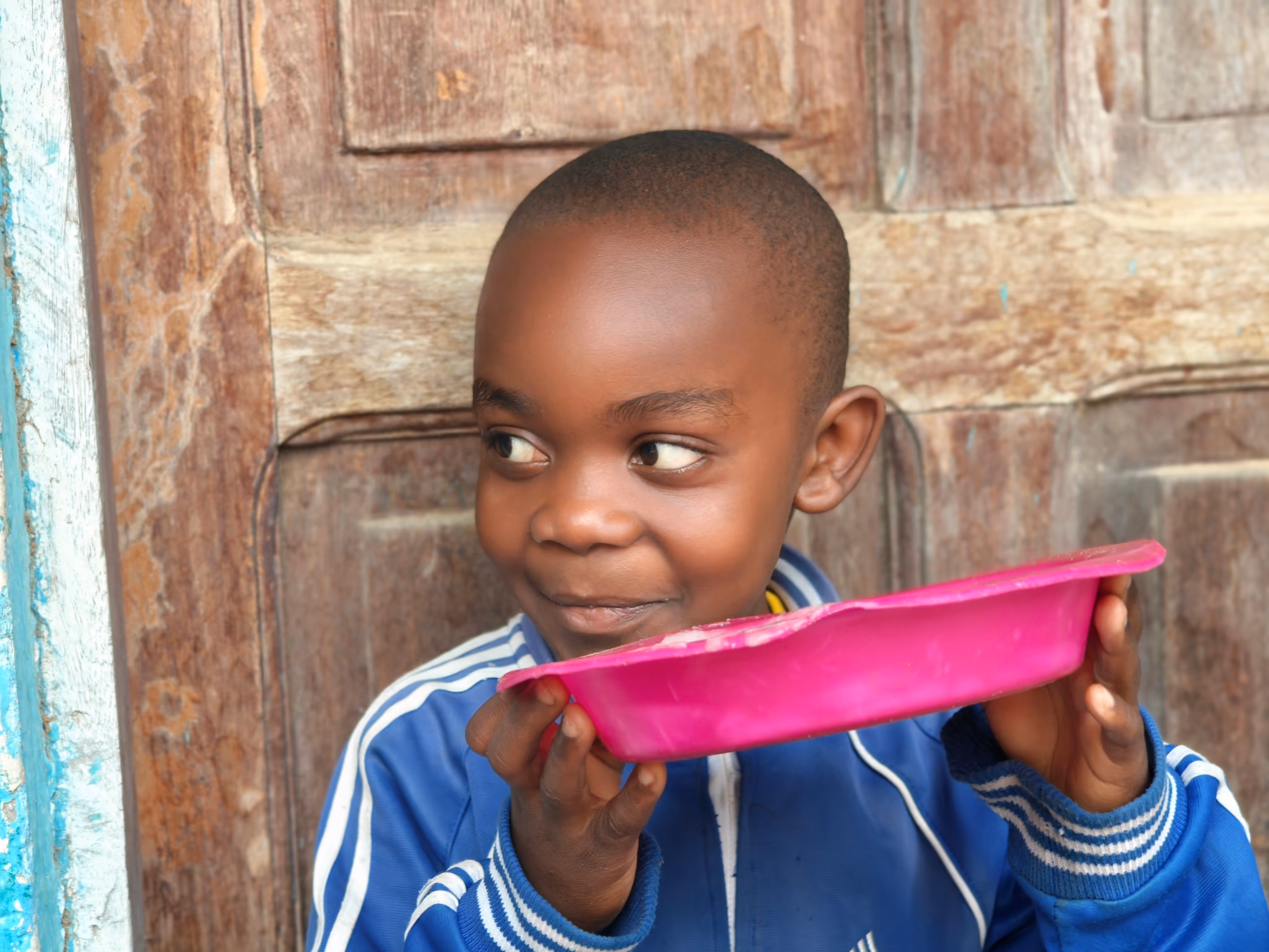 Young boy in a blue jacket holding a pink plate near his mouth with a playful expression.