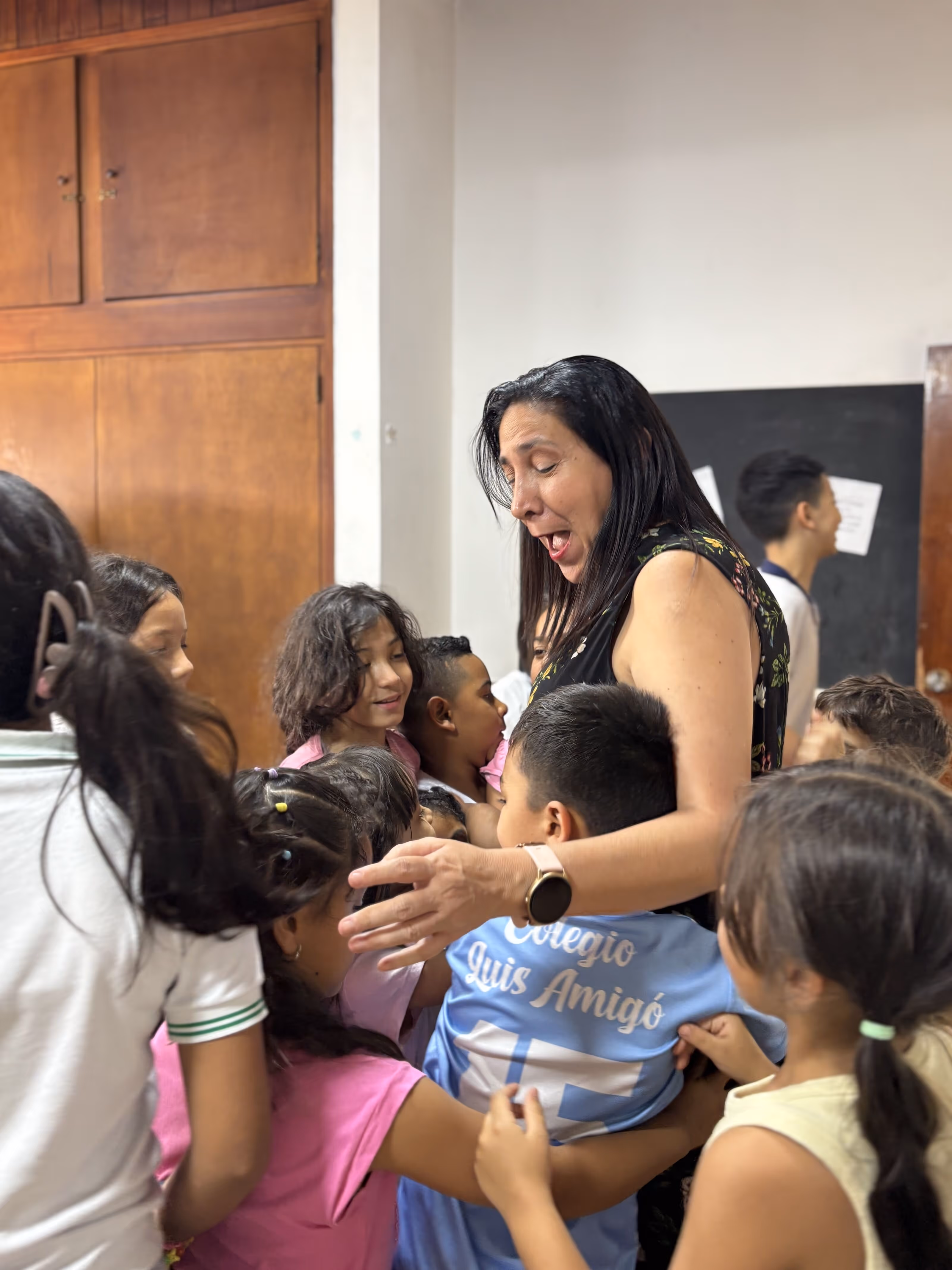 Woman surrounded by children who are hugging her warmly in a classroom.