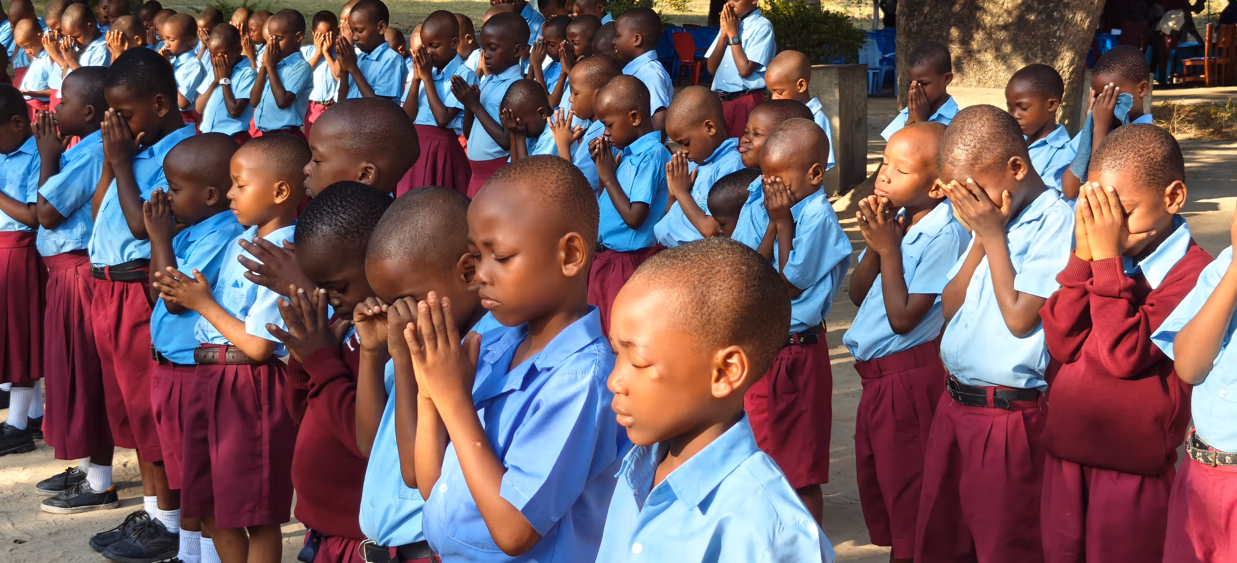Group of young African school children in blue shirts and maroon bottoms standing outdoors with eyes closed and hands clasped in prayer.