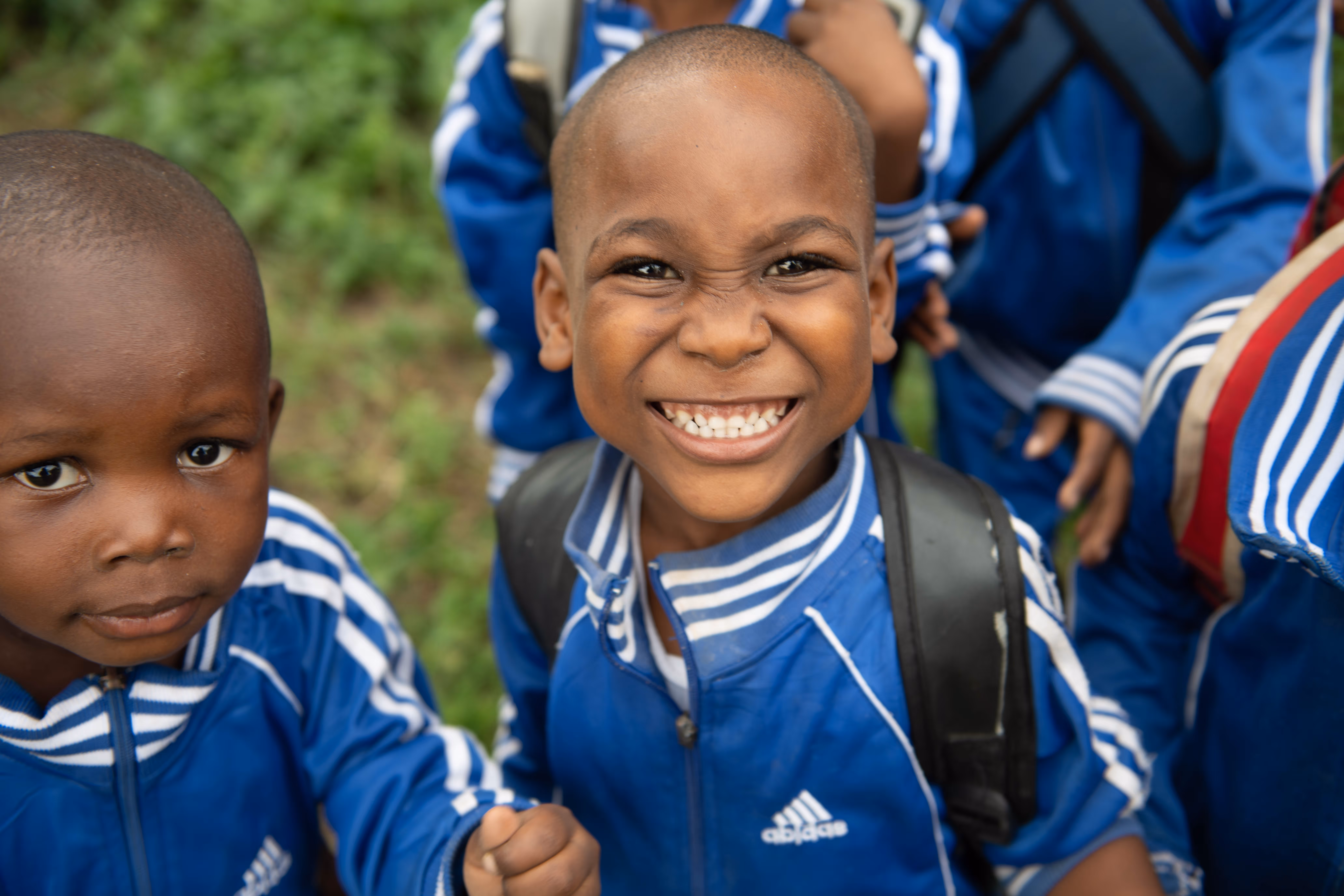 Smiling boy in blue Adidas jacket and backpack with other children in blue jackets around him.