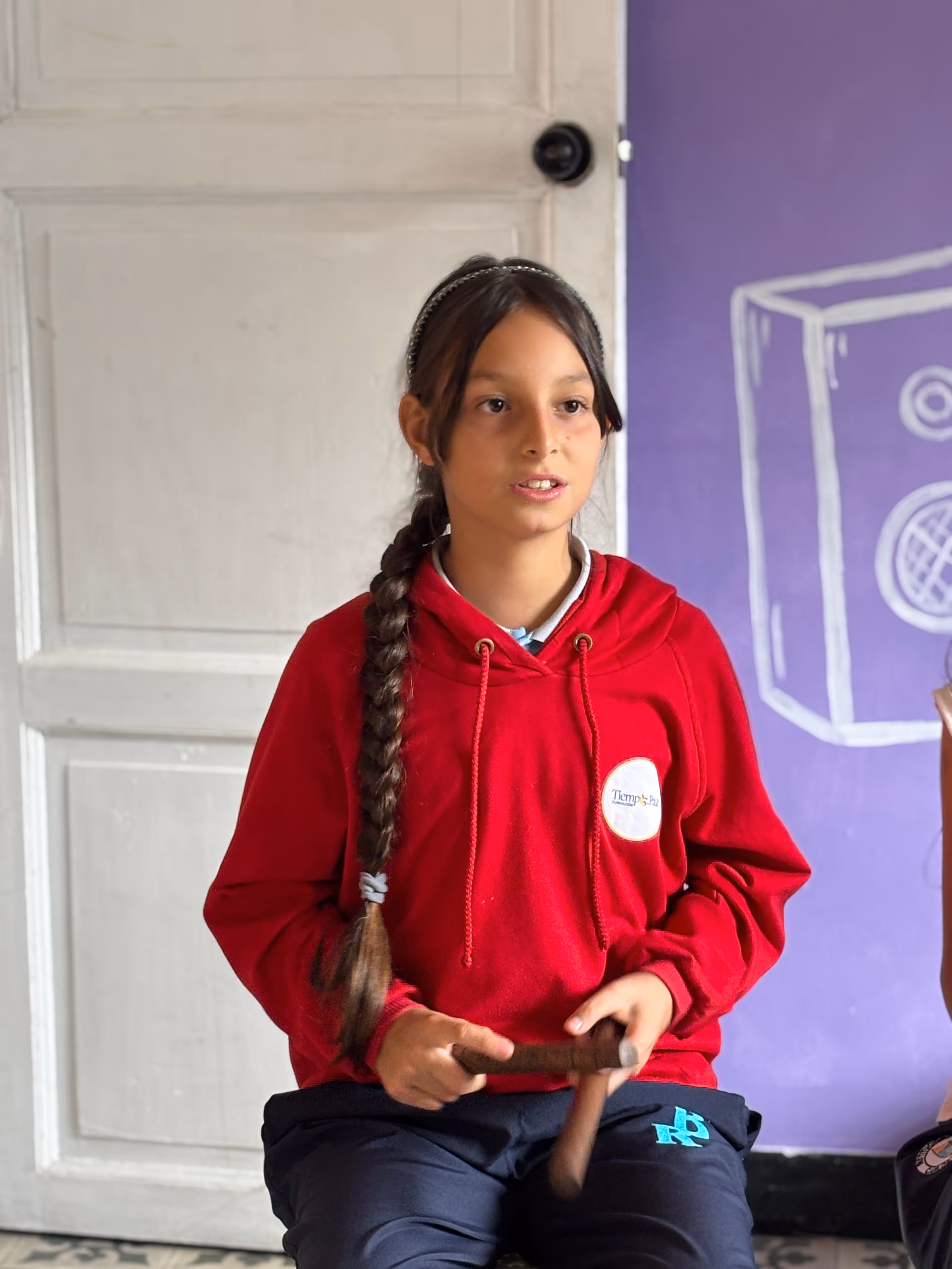 Girl with a long braid wearing a red hoodie playing wooden claves indoors with a purple wall and white door in the background.