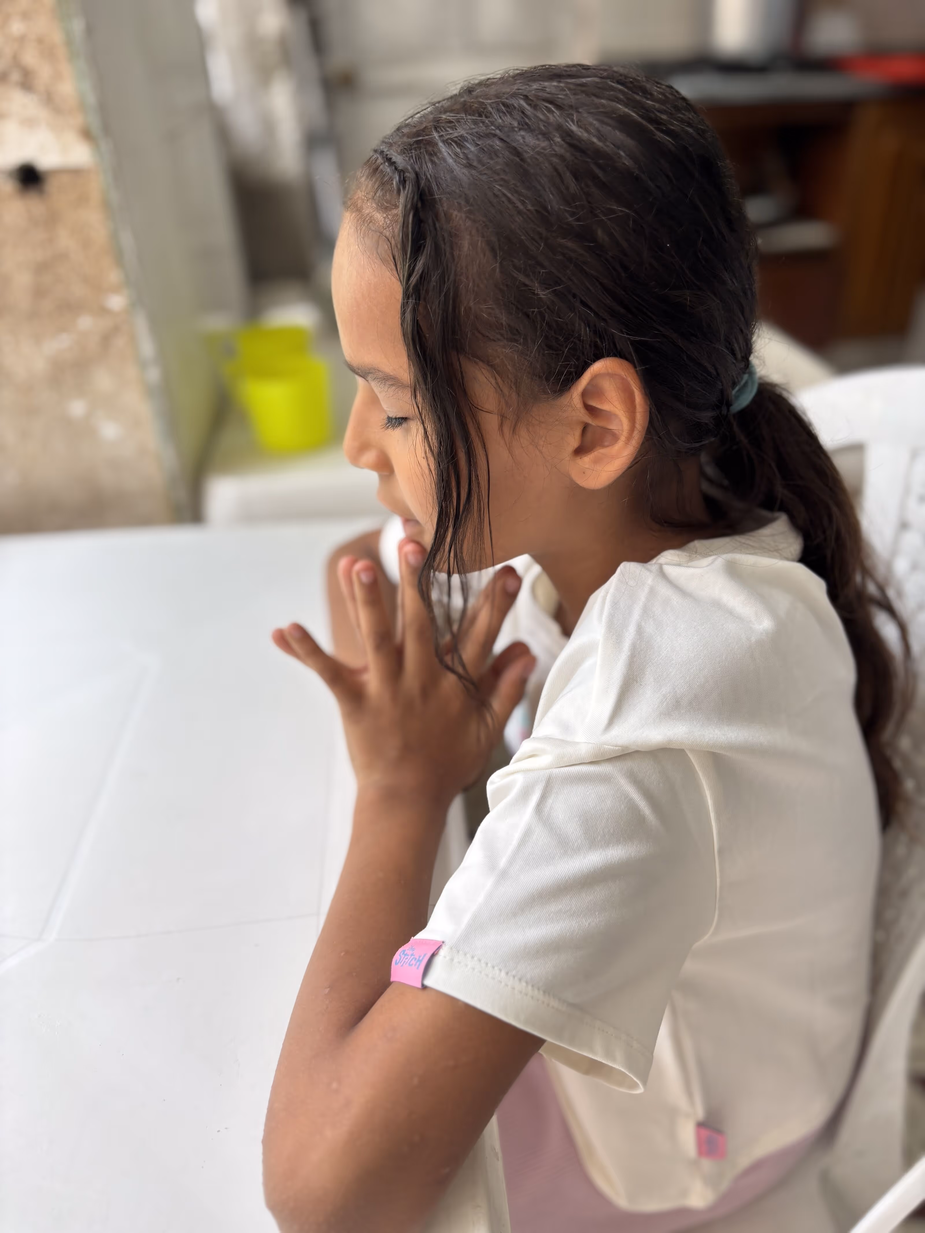Young girl with dark hair tied back sitting at a white table with her hands clasped near her face and eyes closed.