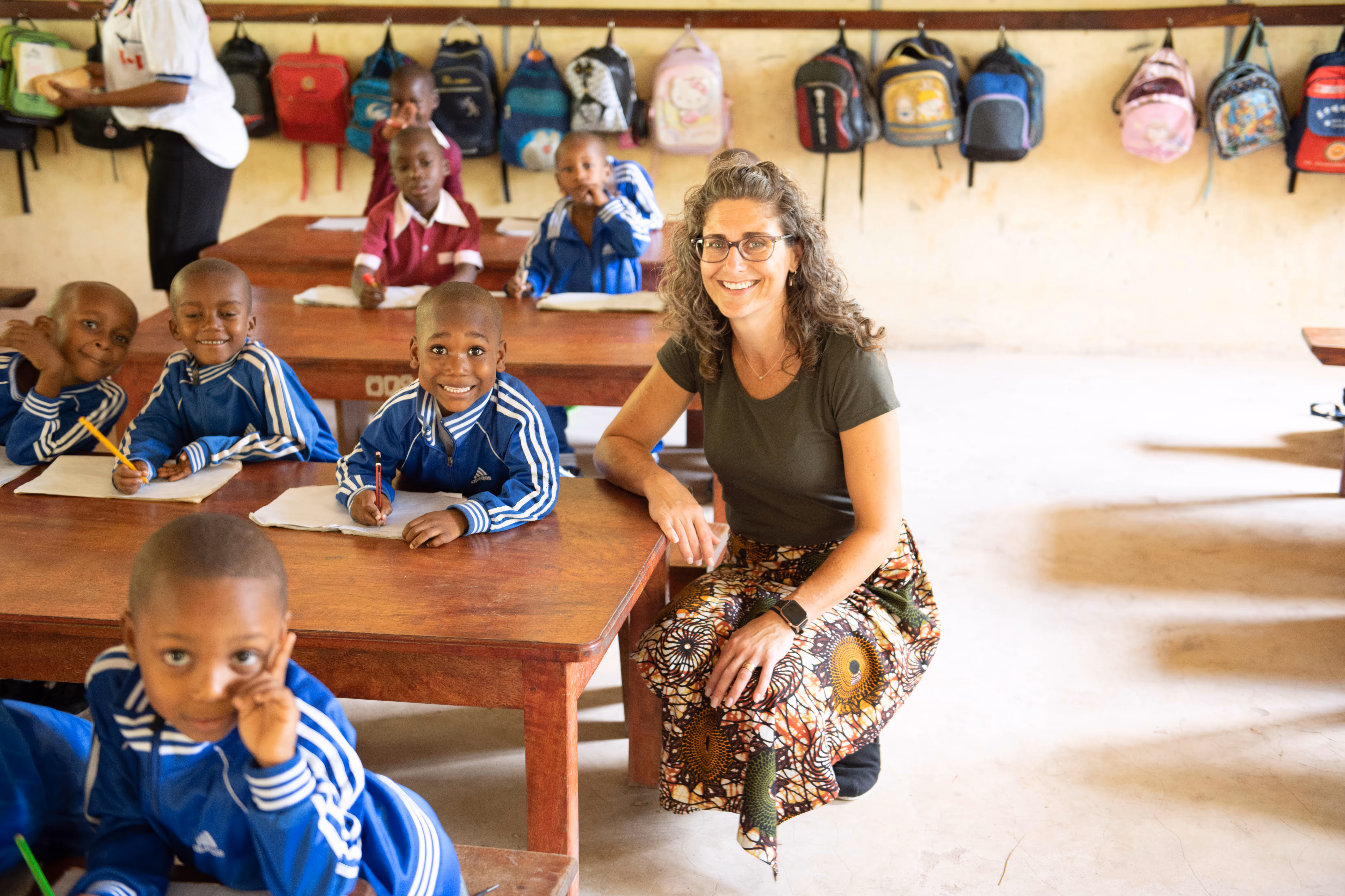 Smiling teacher kneeling beside desks with young students in blue tracksuits writing on paper in a classroom with backpacks hanging on the wall.