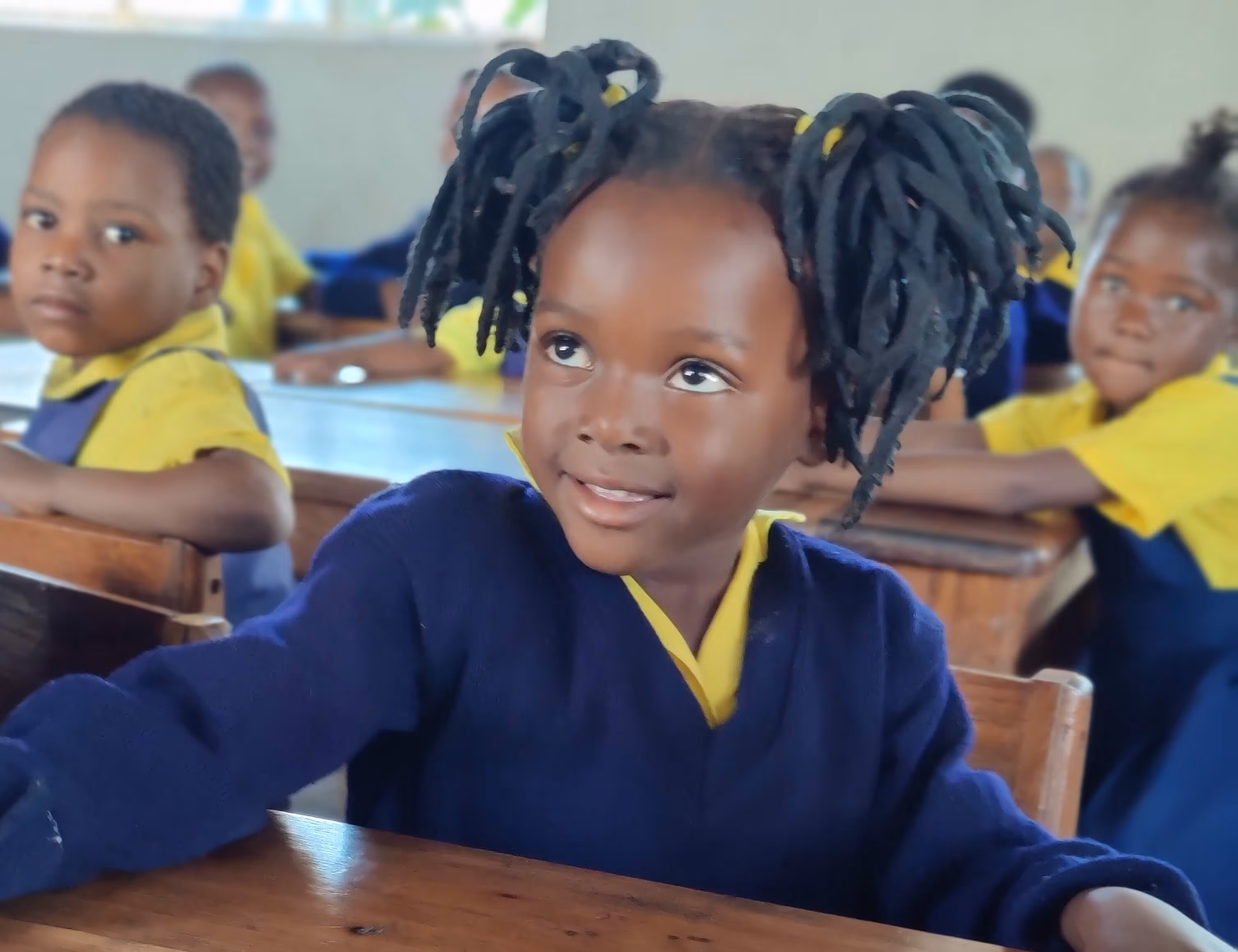 Young schoolgirl with braided pigtails in navy sweater and yellow shirt sitting at a wooden desk in a classroom.