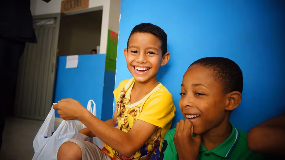 Two boys smiling and sitting against a blue wall, one holding a white plastic bag.