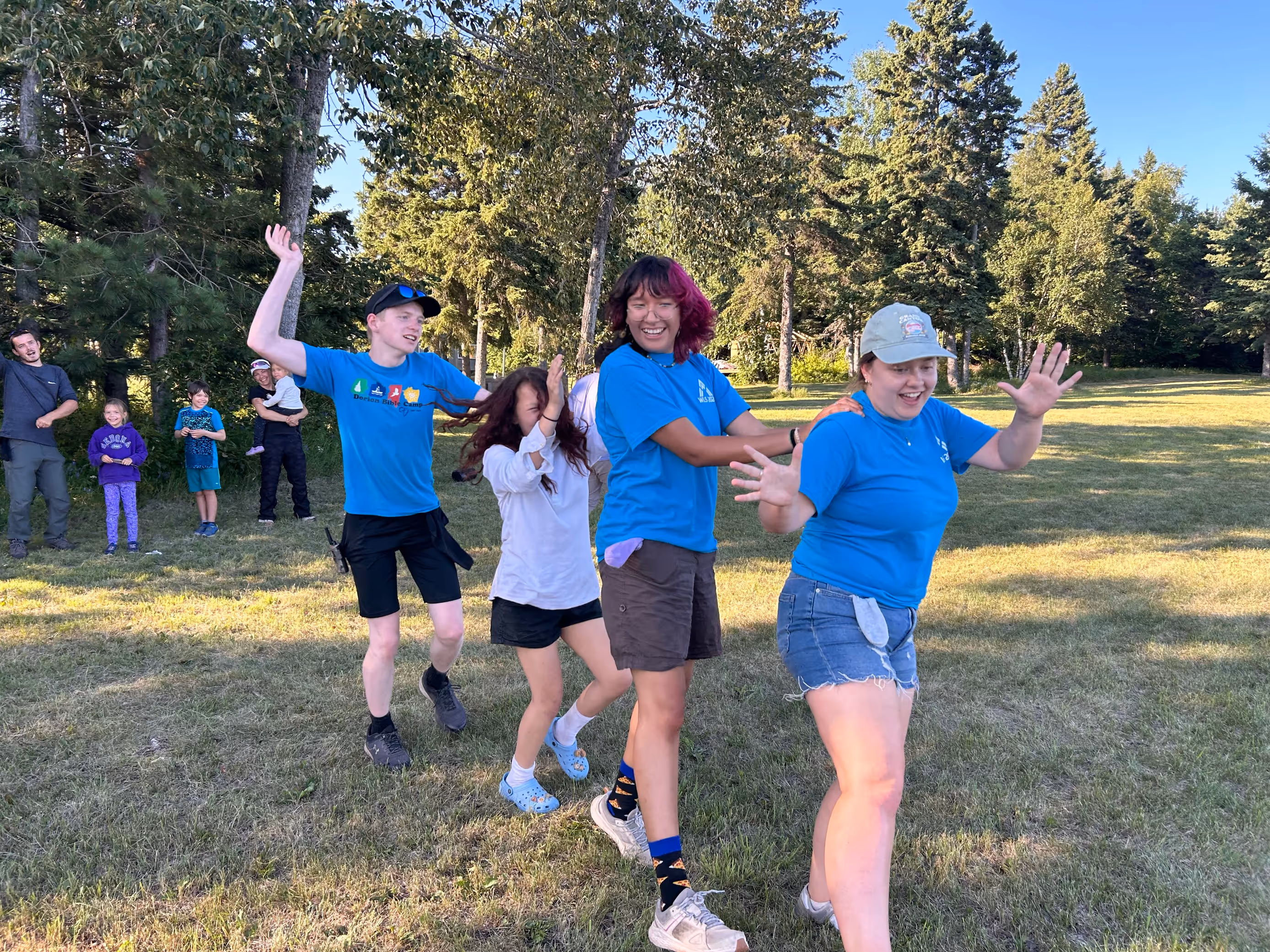 Four young people in a line playing a game outdoors on grass in front of trees, with others watching in the background.