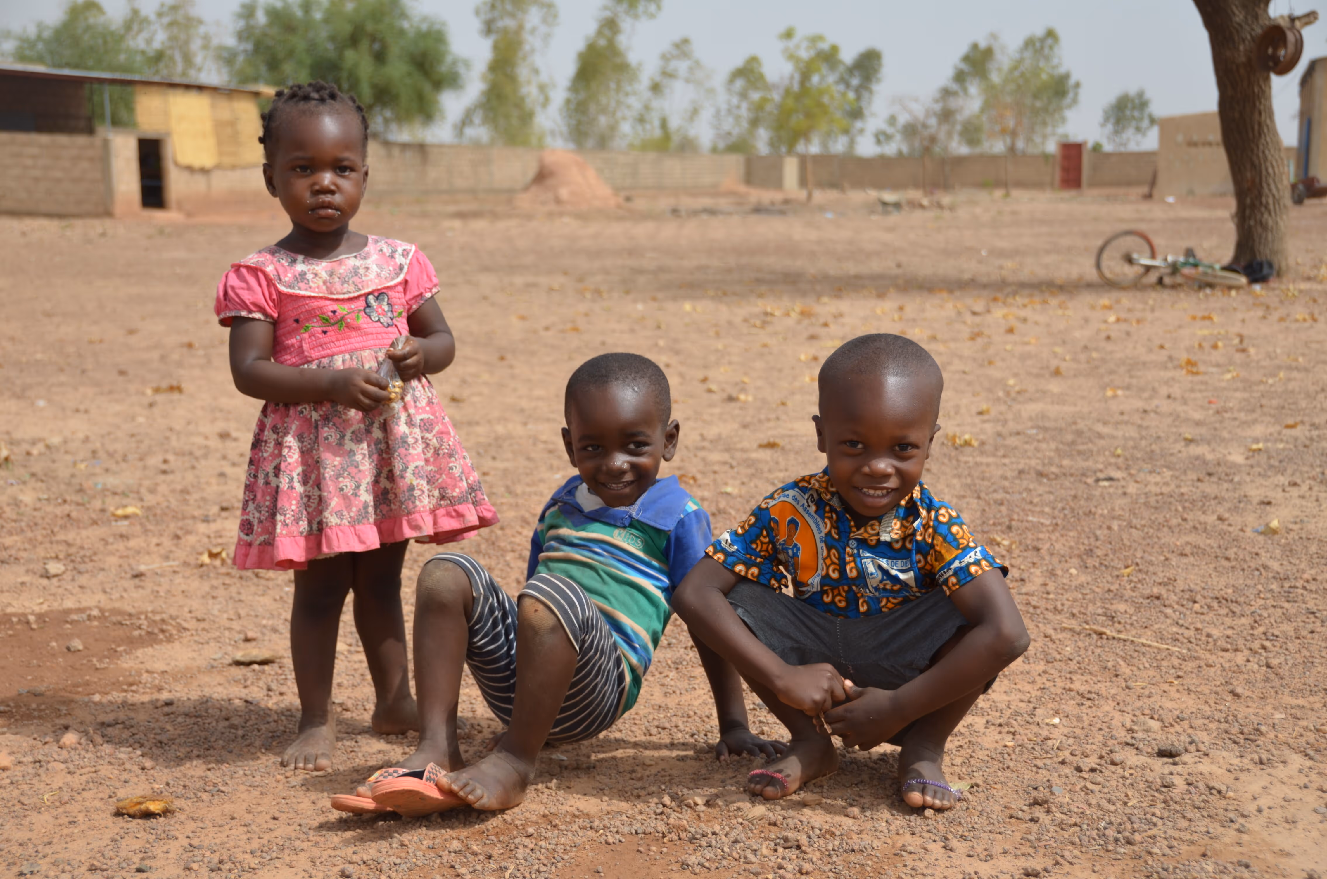 Three young children on a dirt ground outdoors, one standing in a pink dress and two boys sitting and squatting smiling.